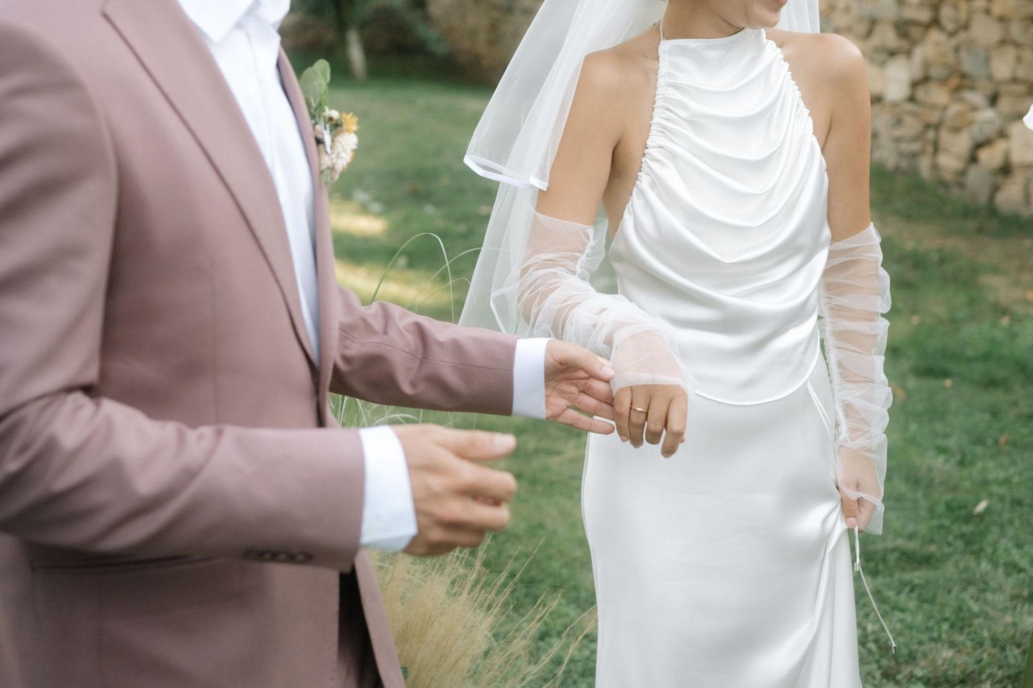 A couple in formal wedding attire clink champagne glasses together during an outdoor celebration.