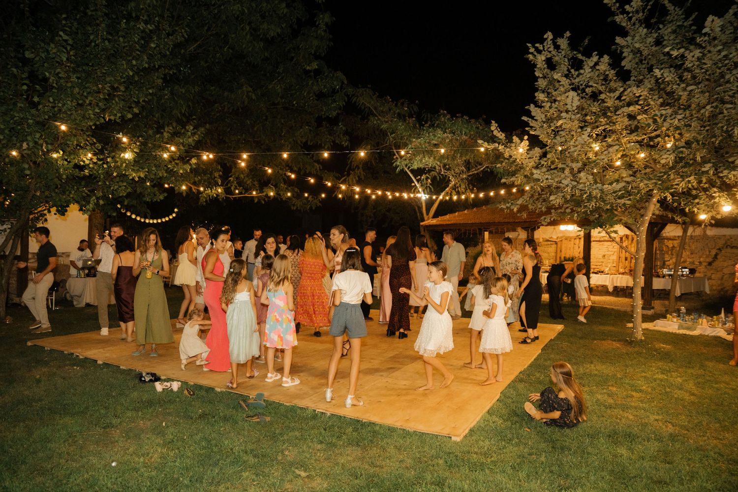 Outdoor evening wedding reception with string lights illuminating guests dancing and mingling in a garden setting.