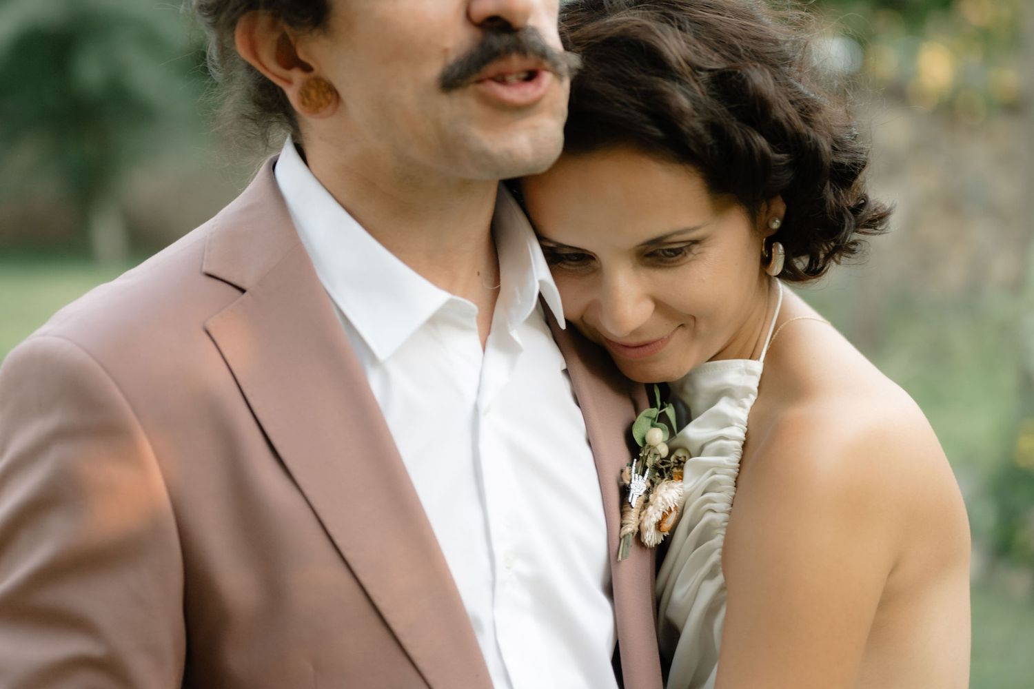 A couple in wedding attire share an intimate moment outdoors surrounded by greenery and natural light.