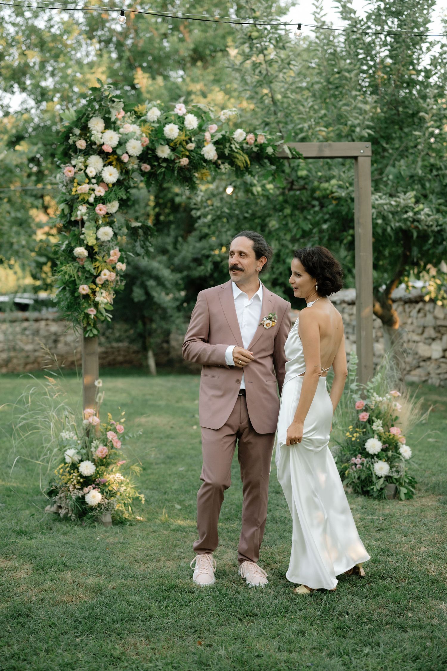 A couple stands beneath a wooden arch decorated with lush white roses and greenery in an outdoor garden setting.