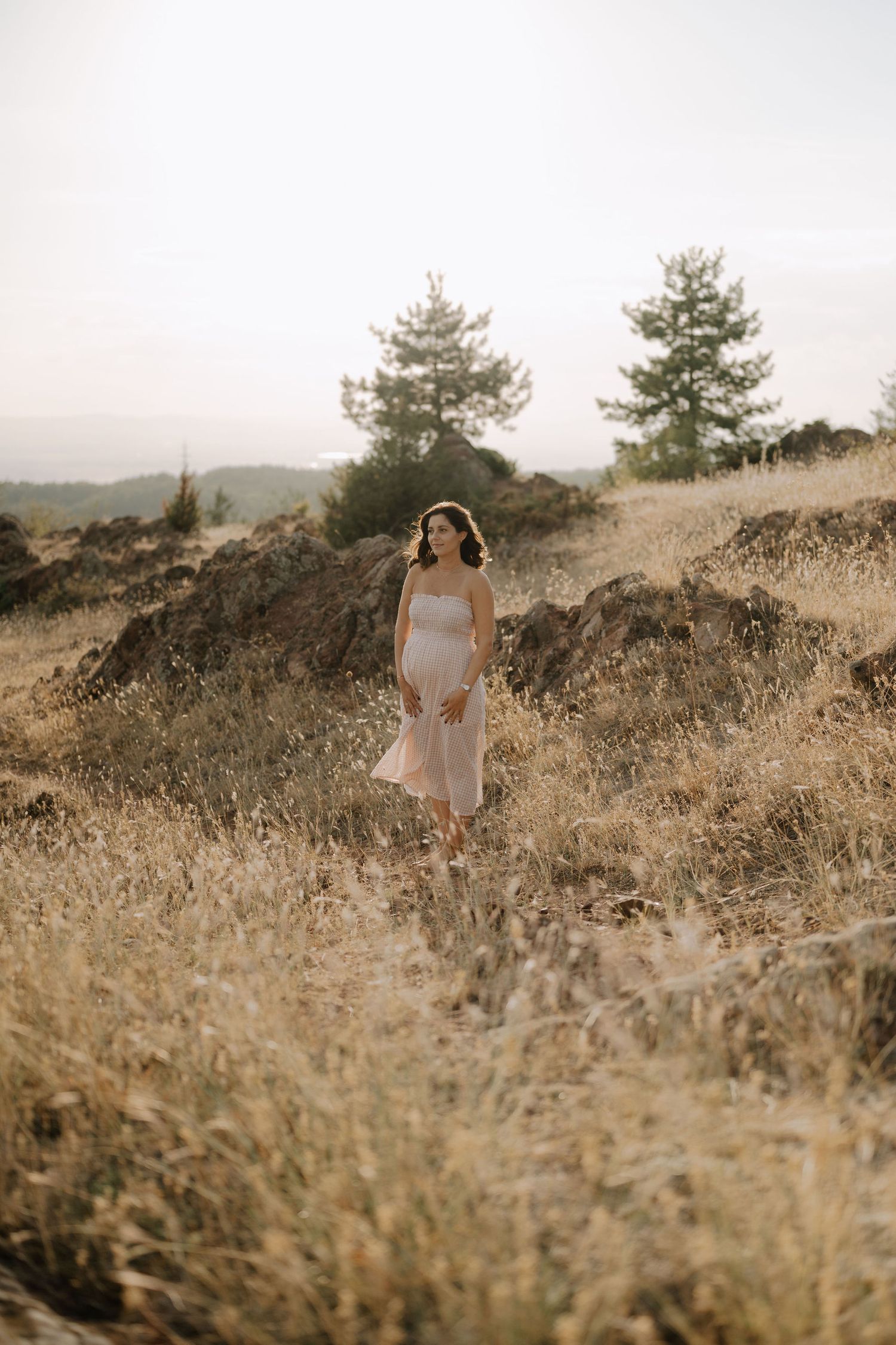 A person in a white dress walks through golden grass on a hillside during sunset with pine trees in the background.