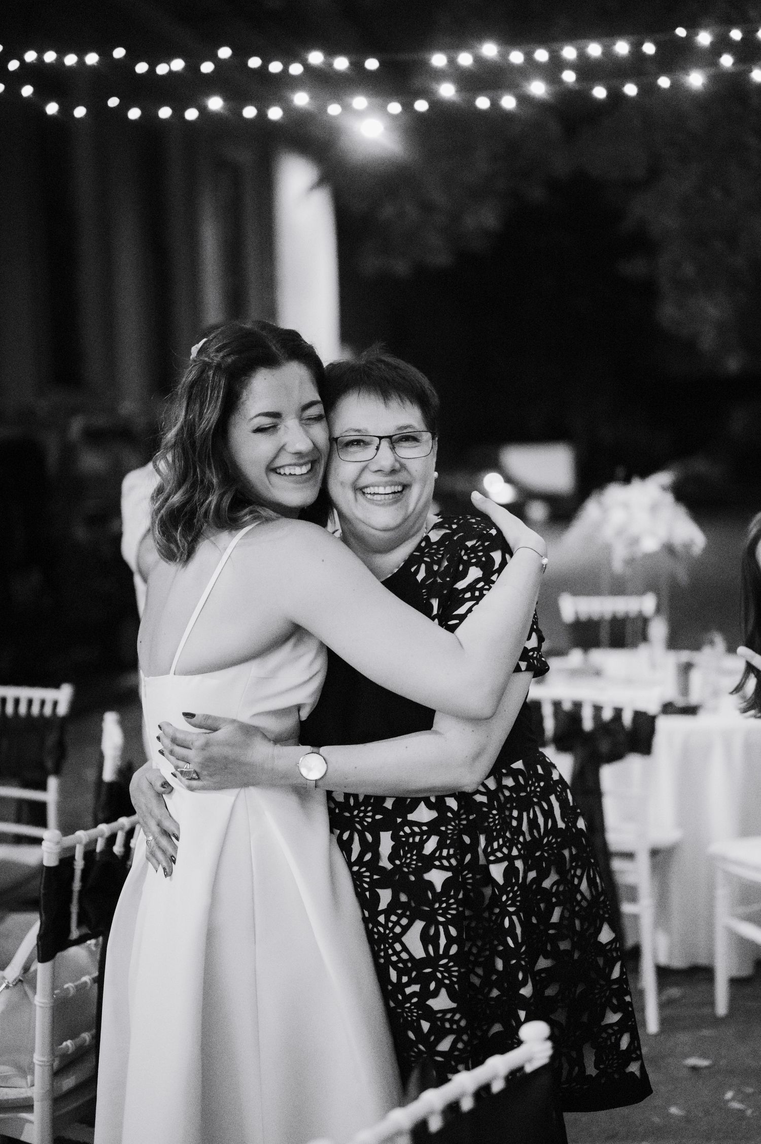 A tender moment captured at a wedding reception with string lights glowing overhead as guests share warm embraces.