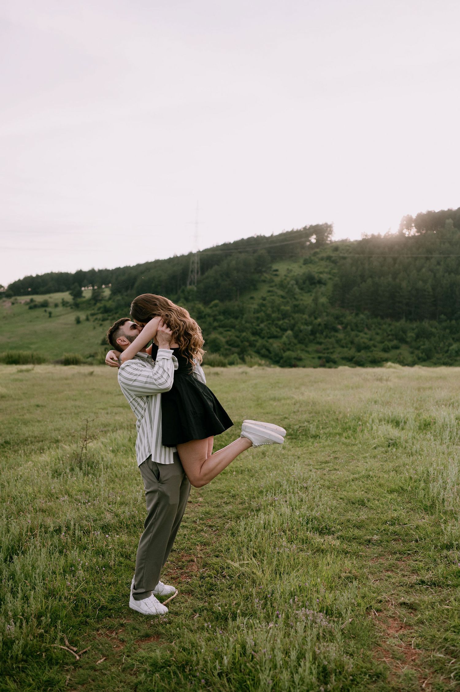 A couple embraces in a romantic moment on a grassy hillside during sunset, with mountains in the background.