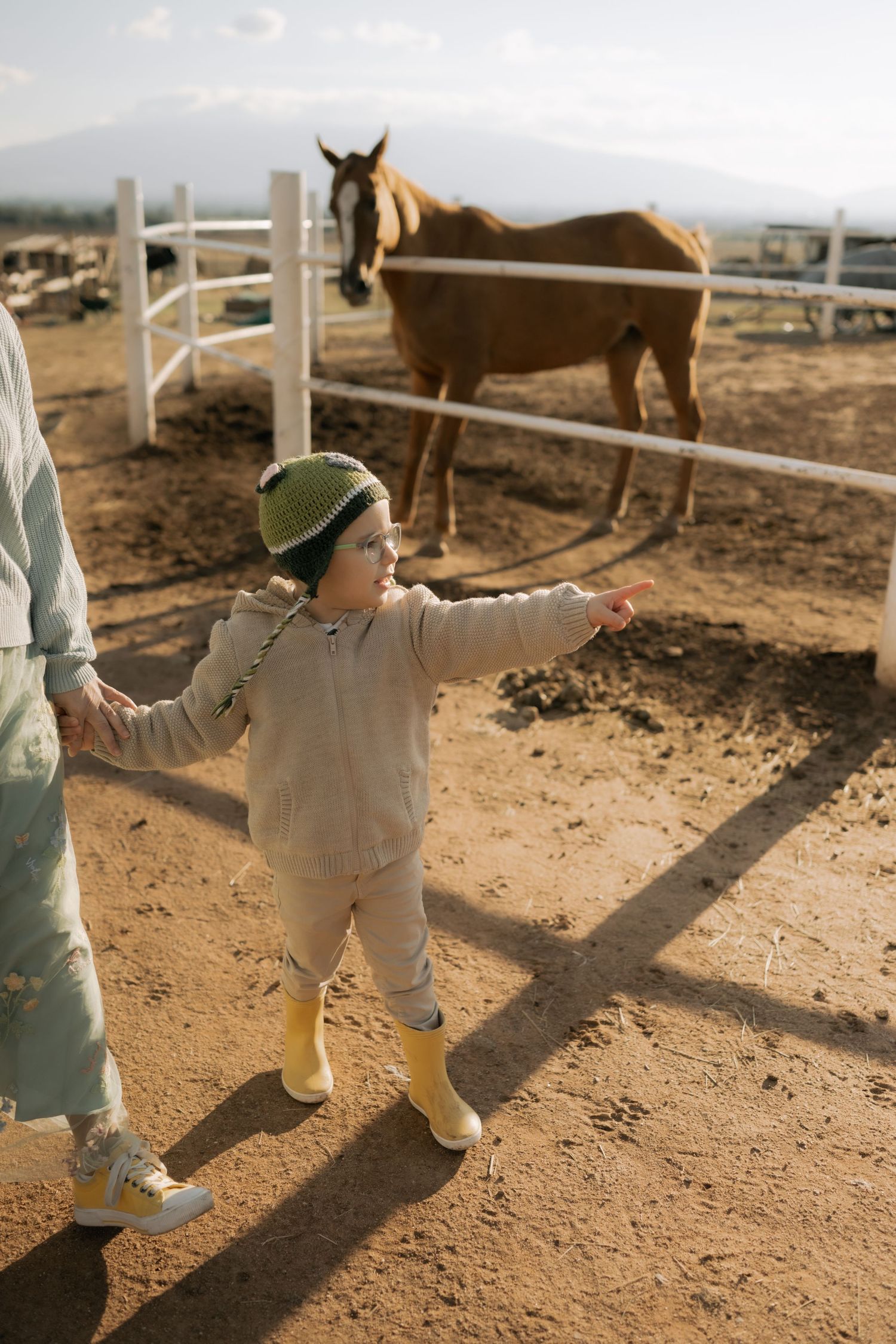 A child in beige clothing plays in a dusty ranch setting with a horse visible in the background.