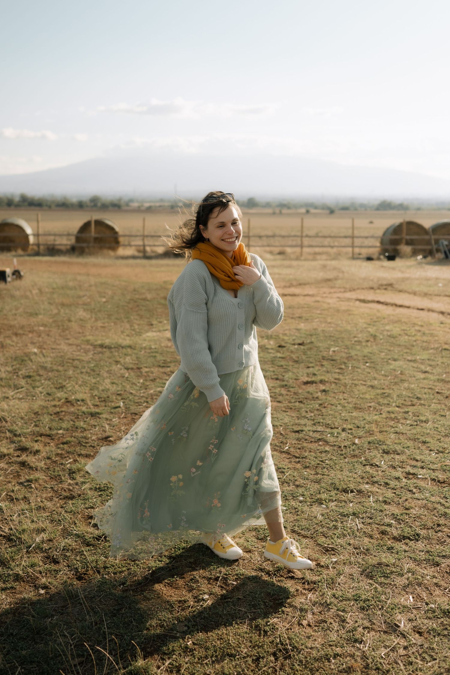 A person in a flowy light blue dress twirls playfully in a golden field at sunset with hay bales in the background.