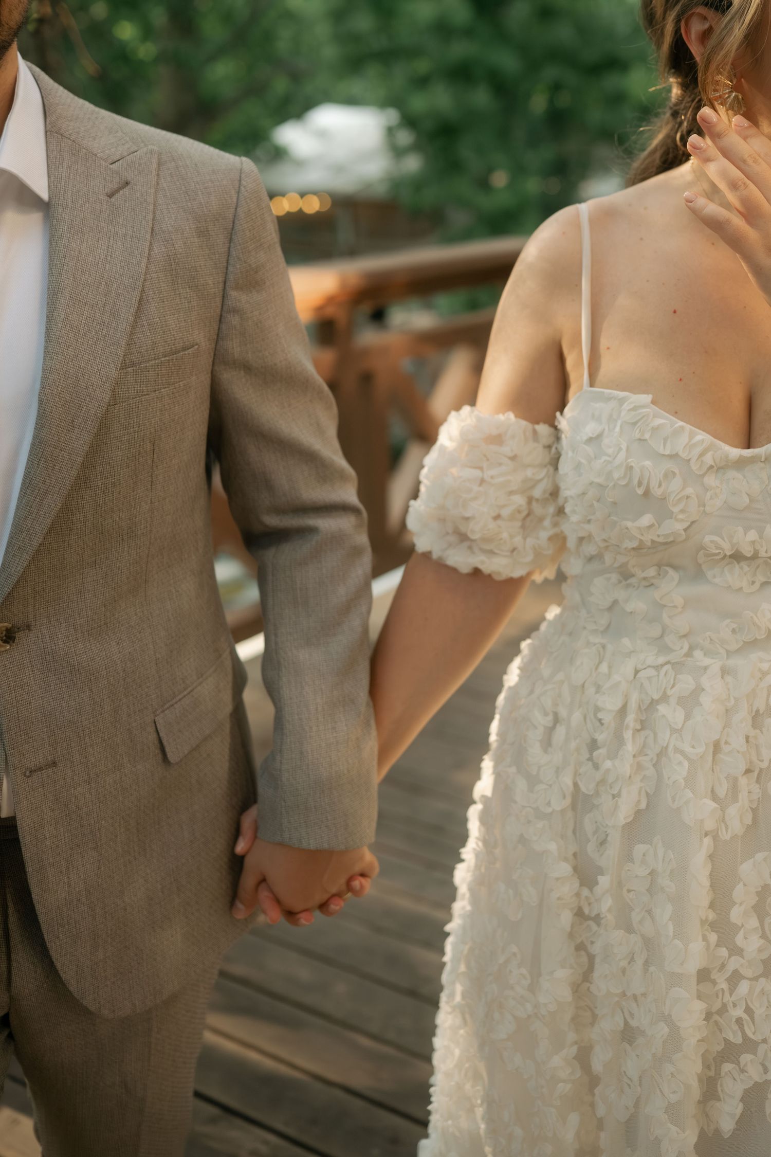 A couple in wedding attire holds hands outdoors, with the bride in a textured white dress and groom in a tan suit.