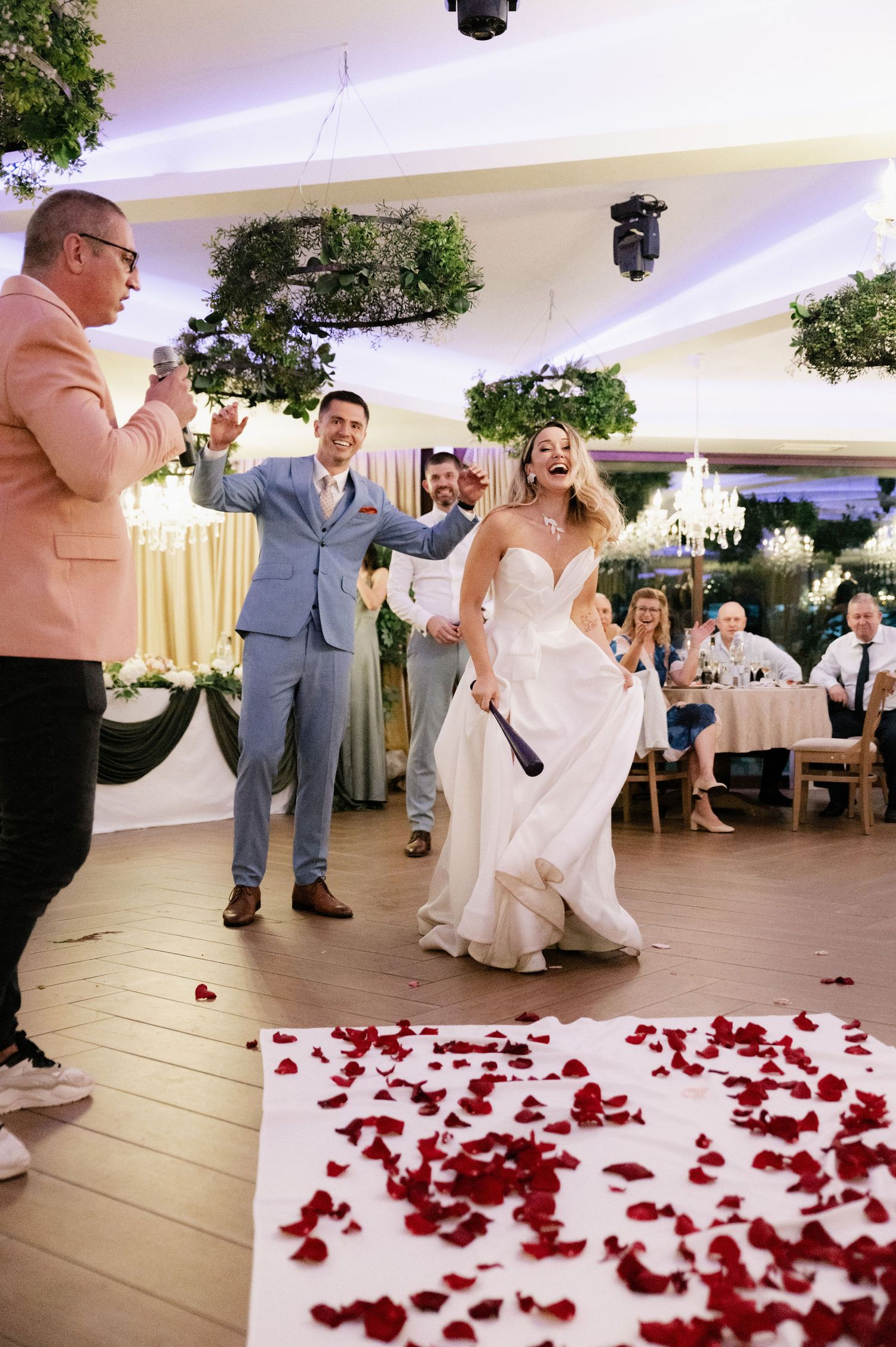 Wedding guests dance and celebrate on a white dance floor decorated with scattered red rose petals at an indoor reception.