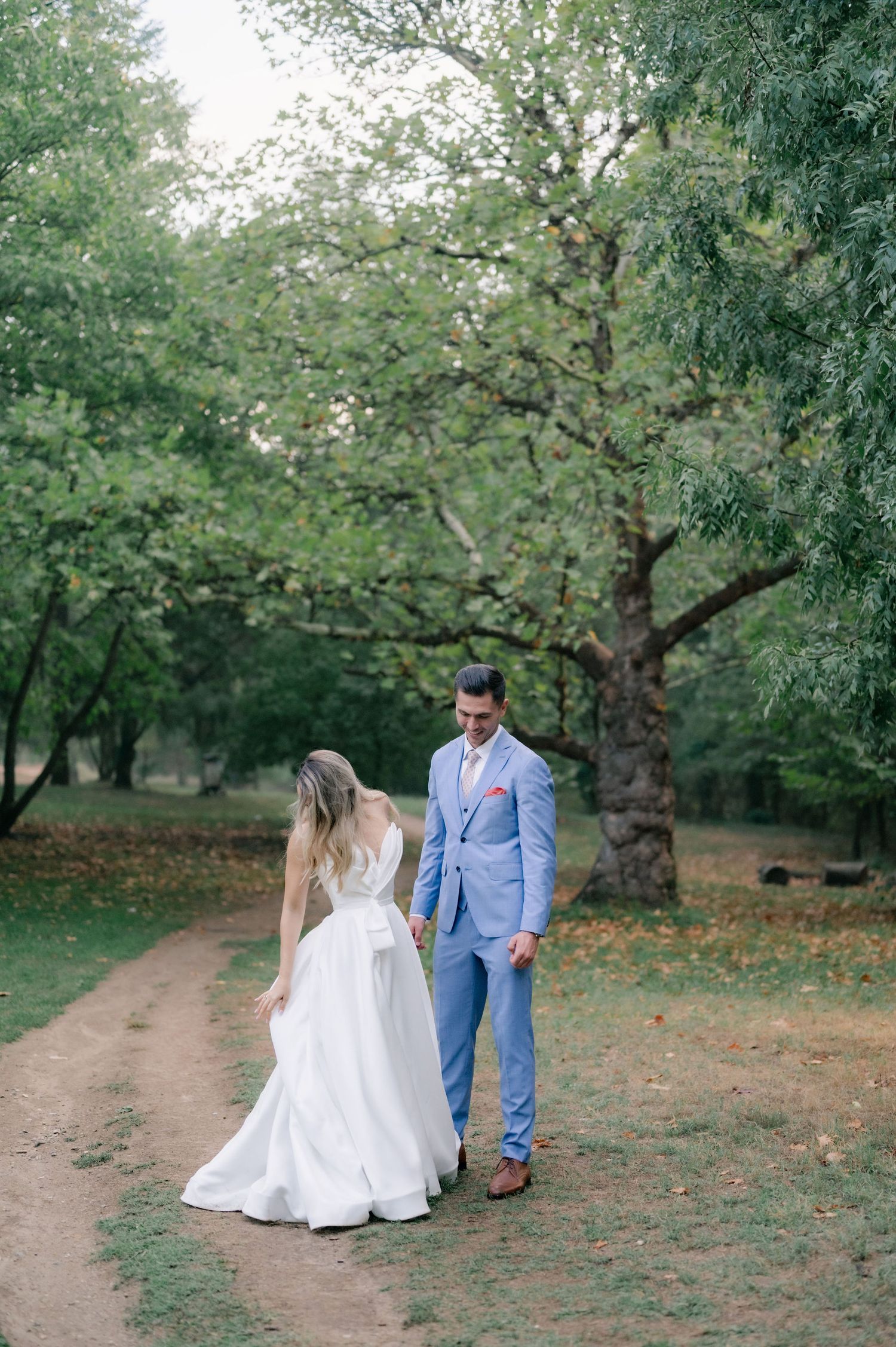 A couple stands together on a rustic dirt path surrounded by flowering trees in a peaceful orchard setting.