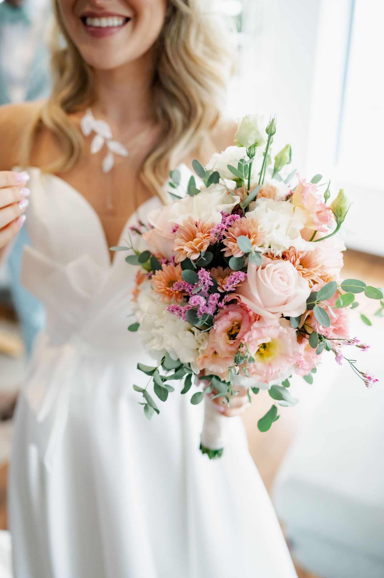 A delicate pastel bouquet with peach roses, pink flowers and eucalyptus held against a white dress in natural light.
