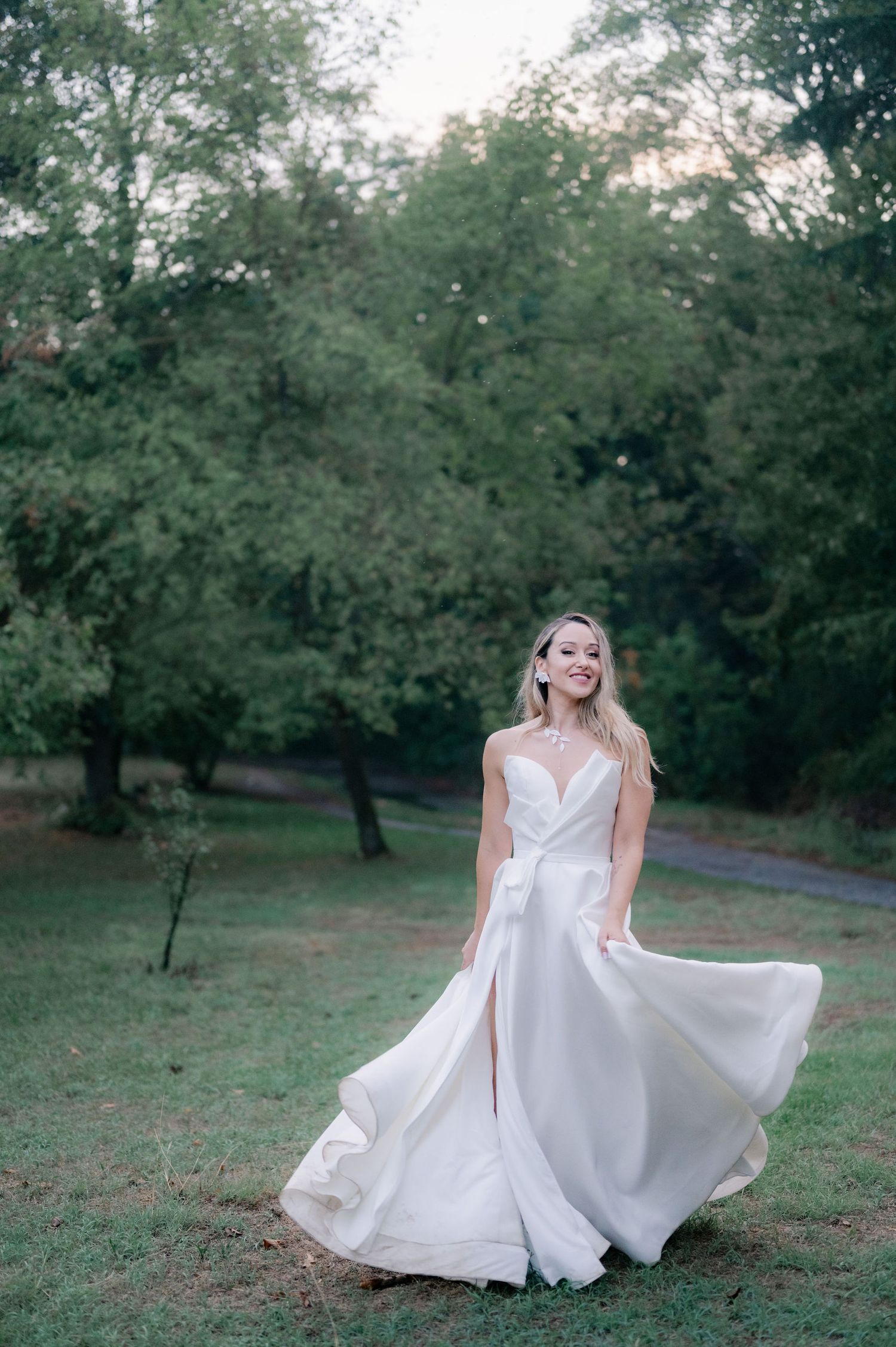 A bride in a flowing white wedding dress twirls gracefully in a scenic outdoor garden setting at dusk.