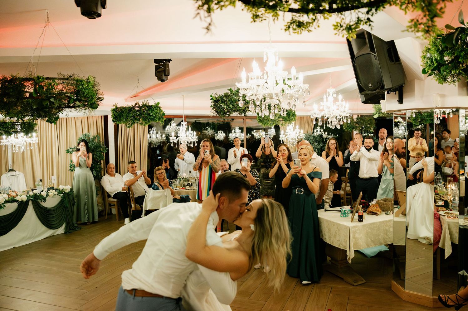 Guests celebrate and dance at an elegant wedding reception with crystal chandeliers and white decor in a banquet hall.