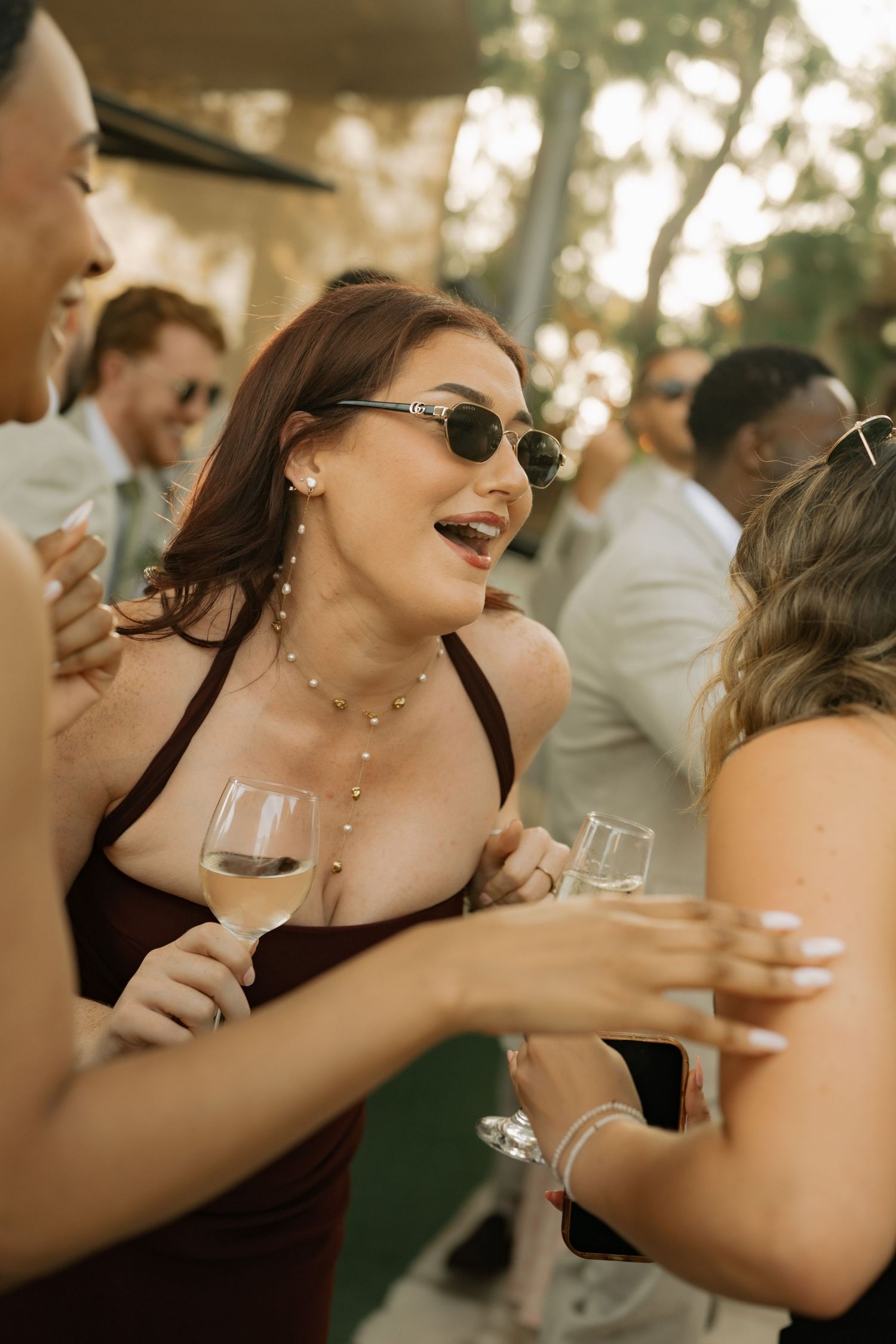 Woman in burgundy dress and sunglasses laughing at outdoor party while holding white wine glass.