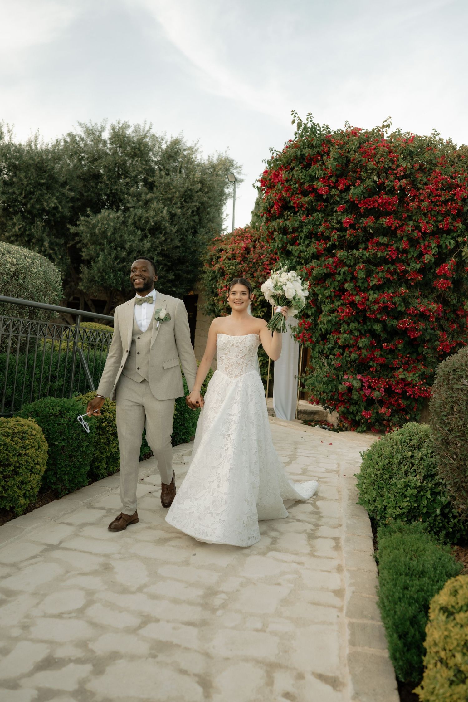 Interracial couple in wedding attire walking hand-in-hand on stone path with red flowering bushes.