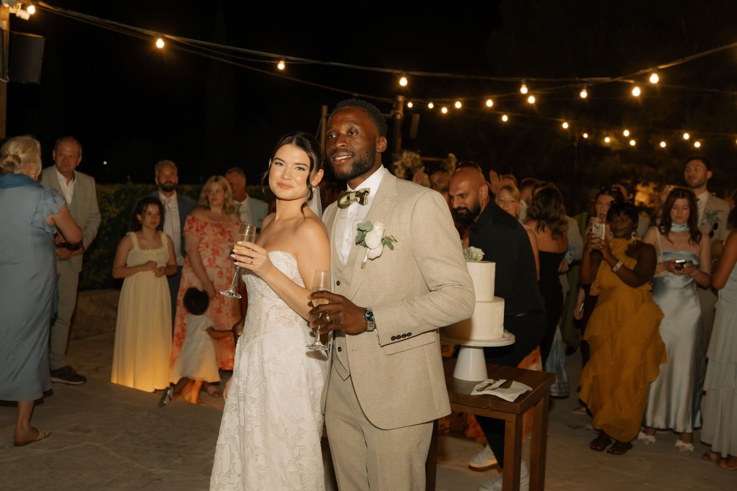 Bride and groom smile together at outdoor evening wedding reception with string lights and guests in background.