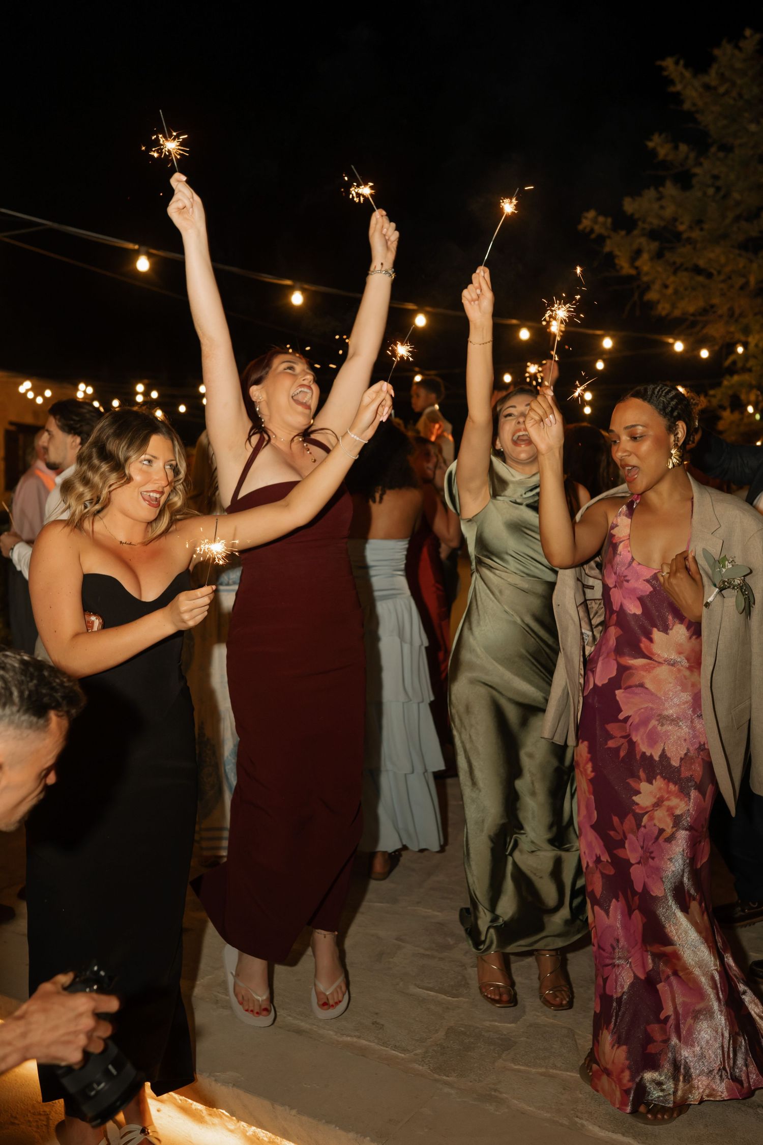 Women in elegant dresses celebrate at a nighttime wedding reception, holding sparklers under string lights.