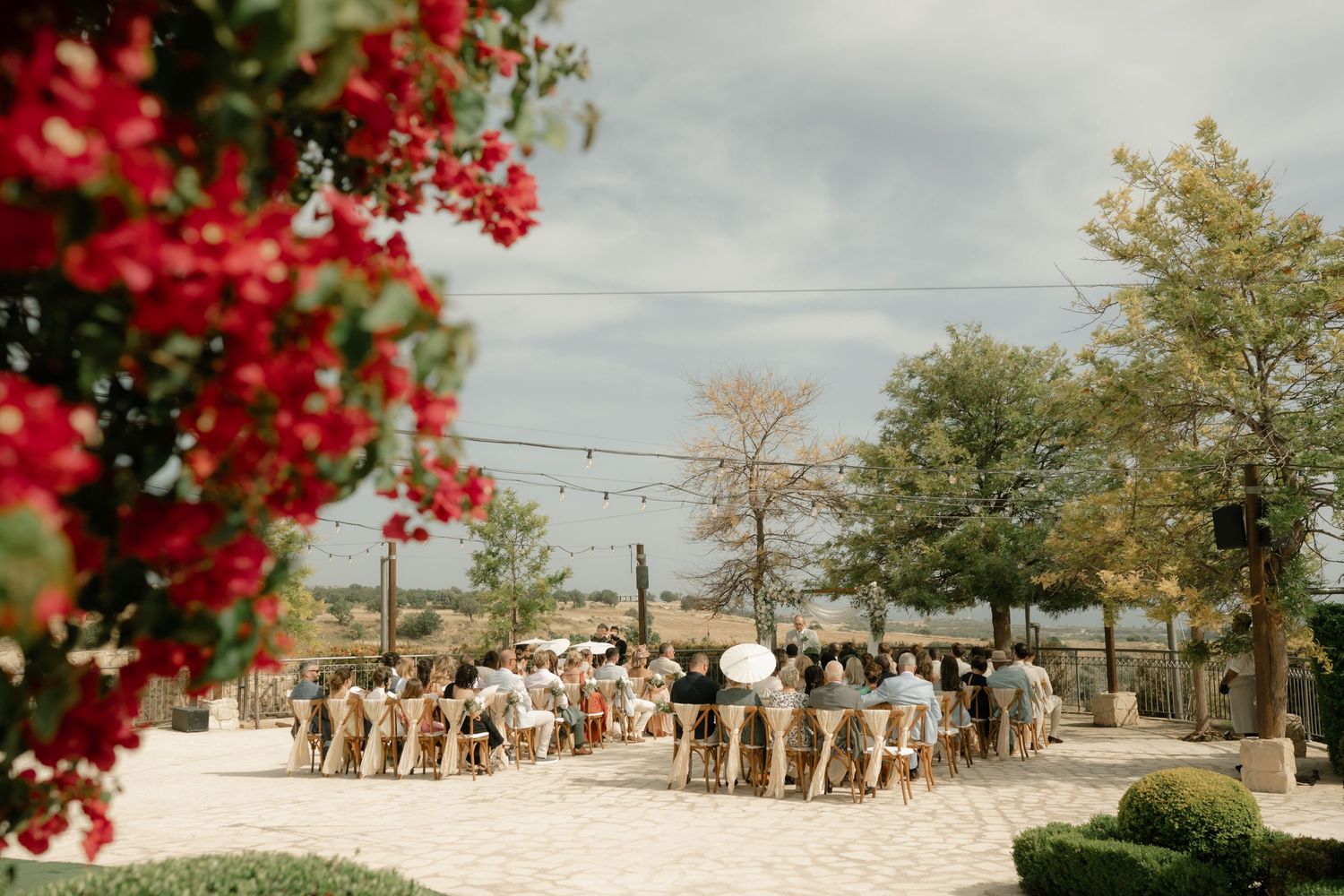 Outdoor wedding ceremony with guests seated on sandy ground, string lights, and red bougainvillea blooms framing the scene.
