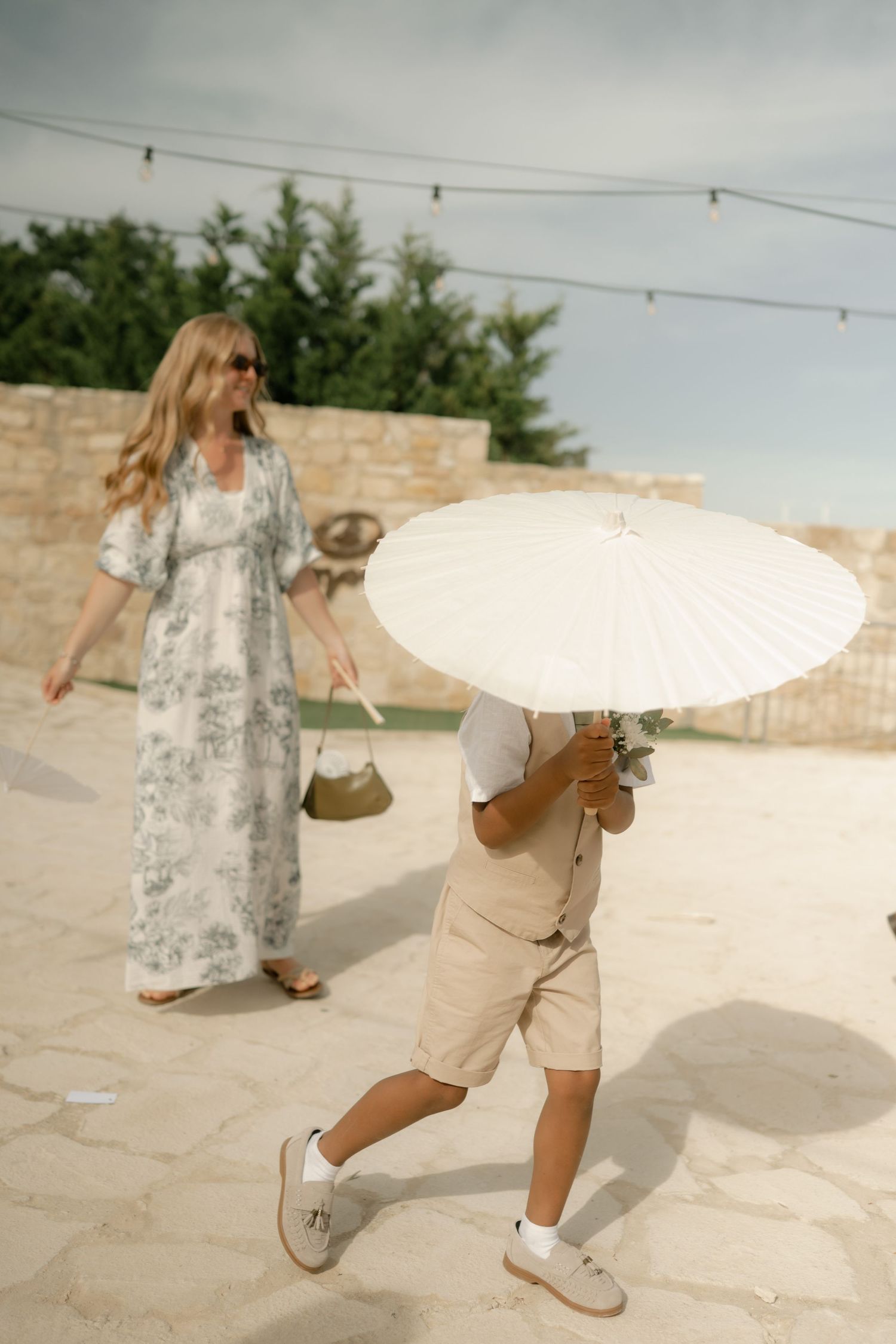 A child holding a white parasol umbrella walks on a stone patio while a woman in a floral maxi dress strolls nearby.