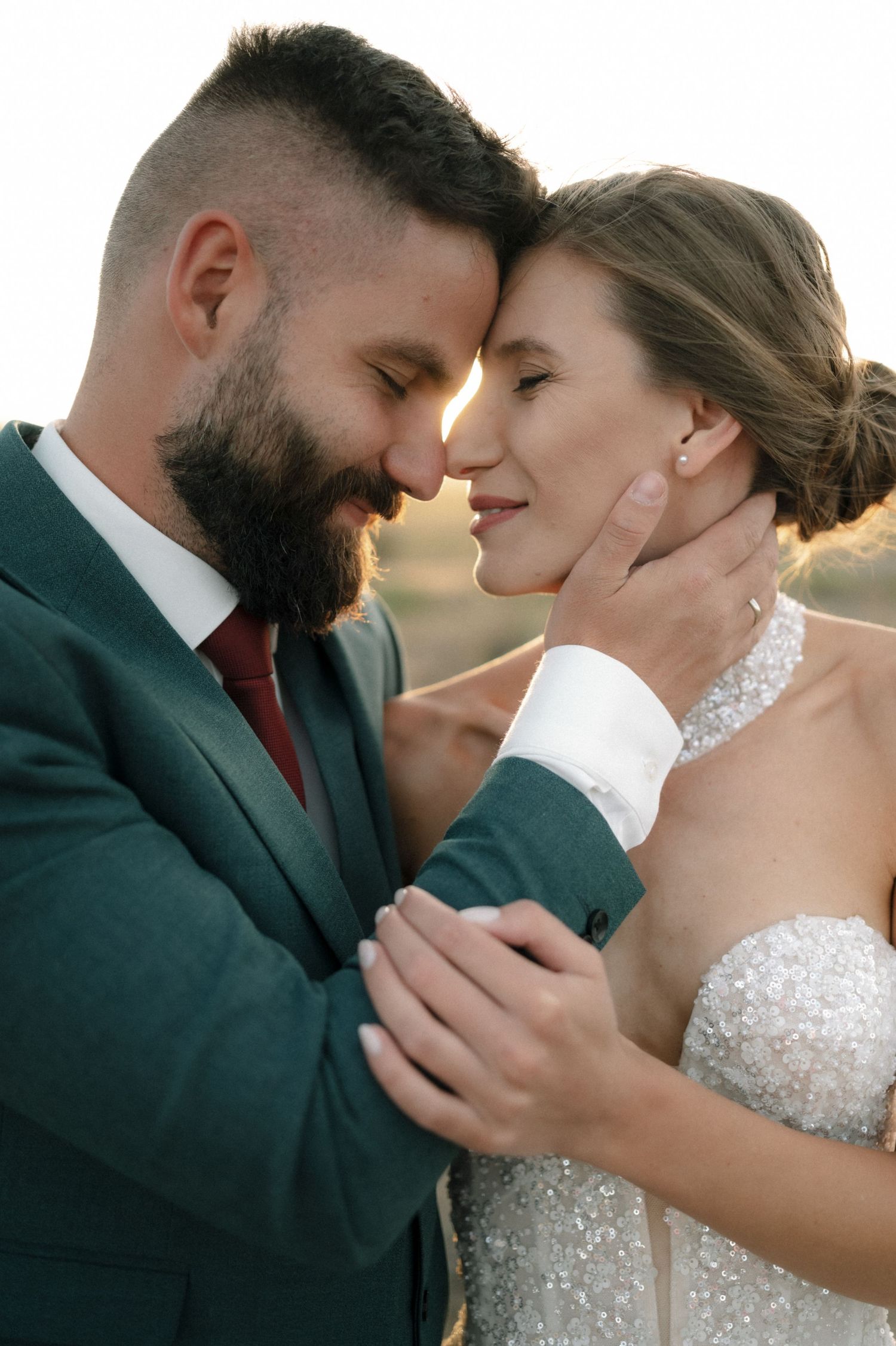 Romantic wedding couple in golden sunset light, groom in green suit with red tie tenderly holding bride in beaded gown.