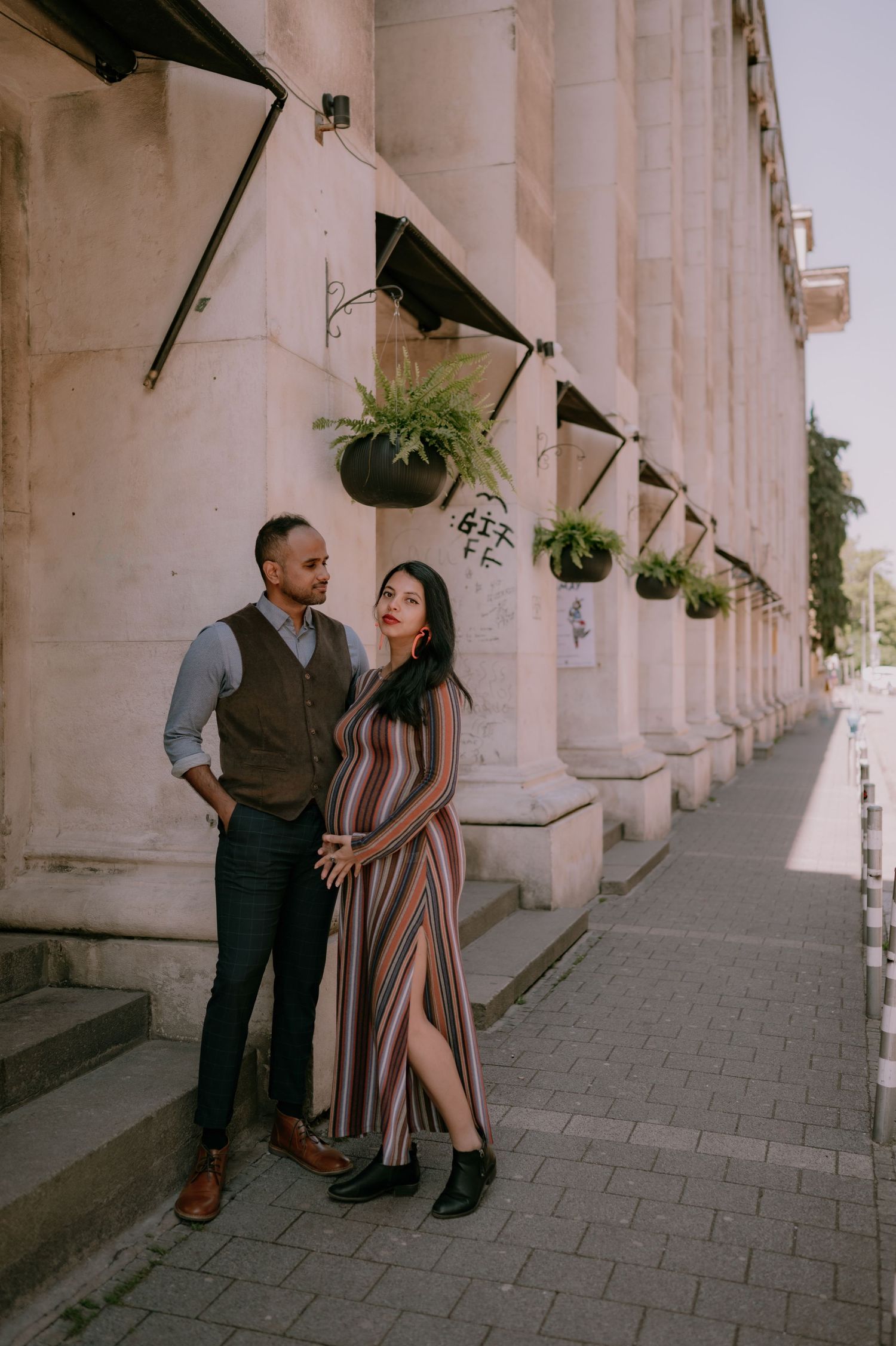 Couple posing by a grand stone building with hanging plants, woman in striped maxi dress, man in vest and trousers.