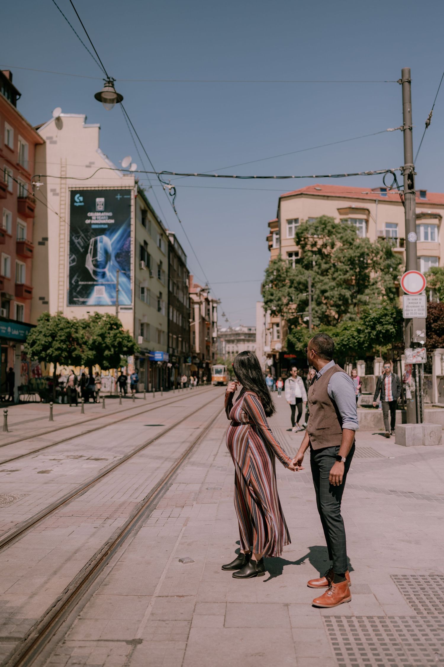 Couple holding hands on a sunny urban street with tram tracks, colorful buildings, and trees in the background.