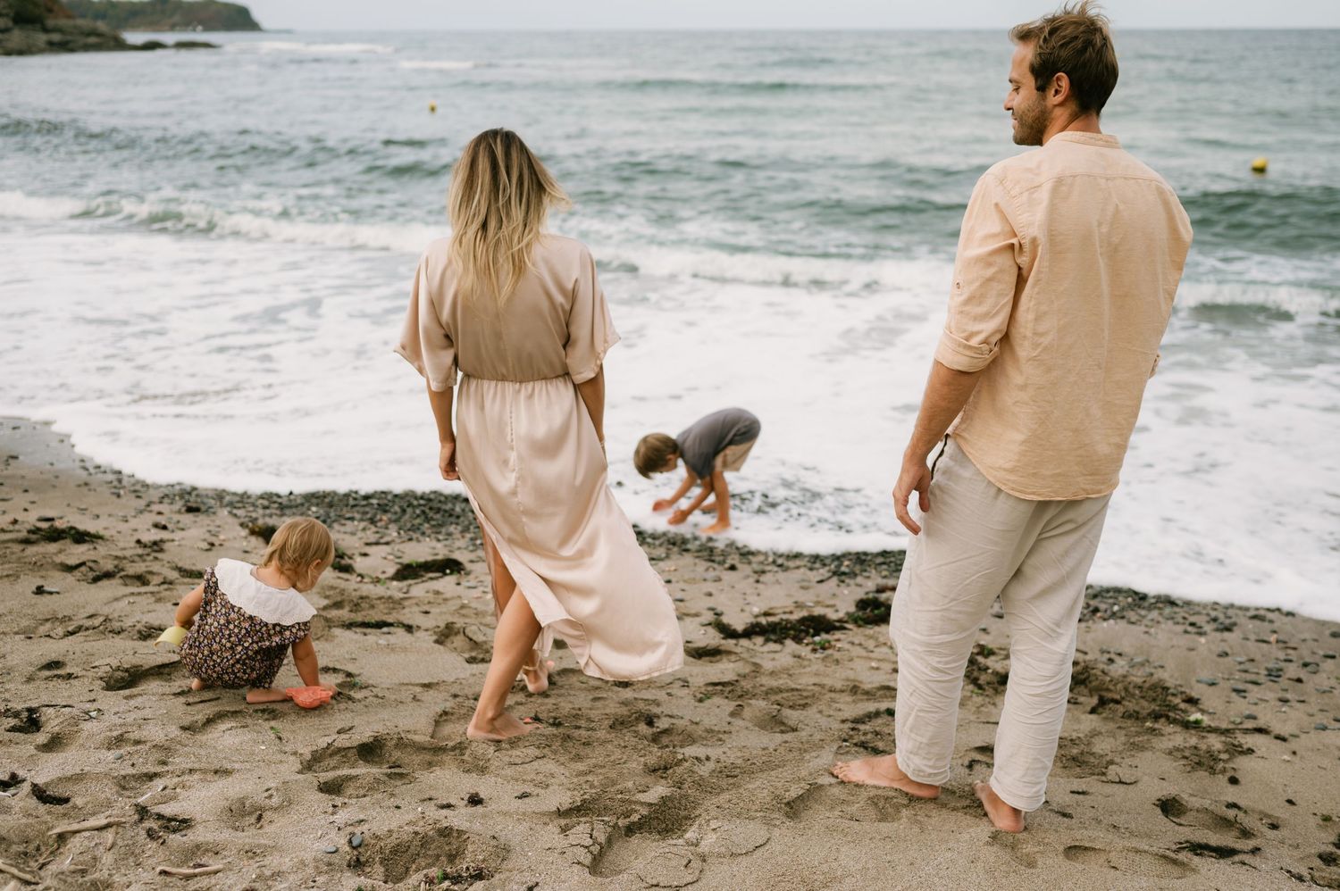 Family of four enjoying a beach day, parents watching children play on sandy shore with ocean waves in background.