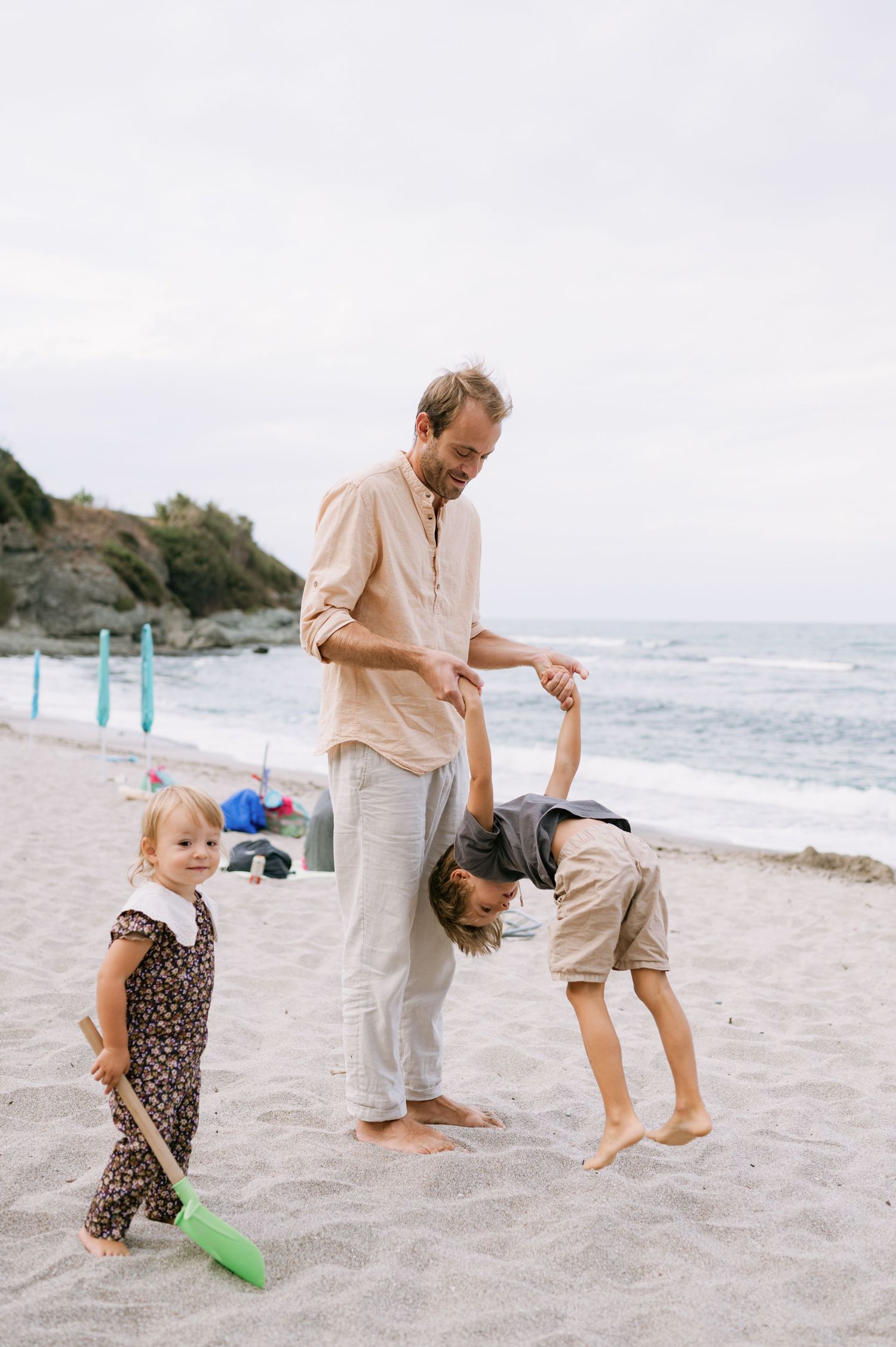 Father plays with two young children on a sandy beach, helping one do a backbend while toddler holds a toy shovel.