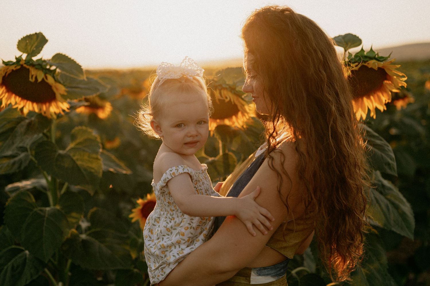 Mother holding toddler in sunflower field at golden hour sunset, warm light glowing behind them.