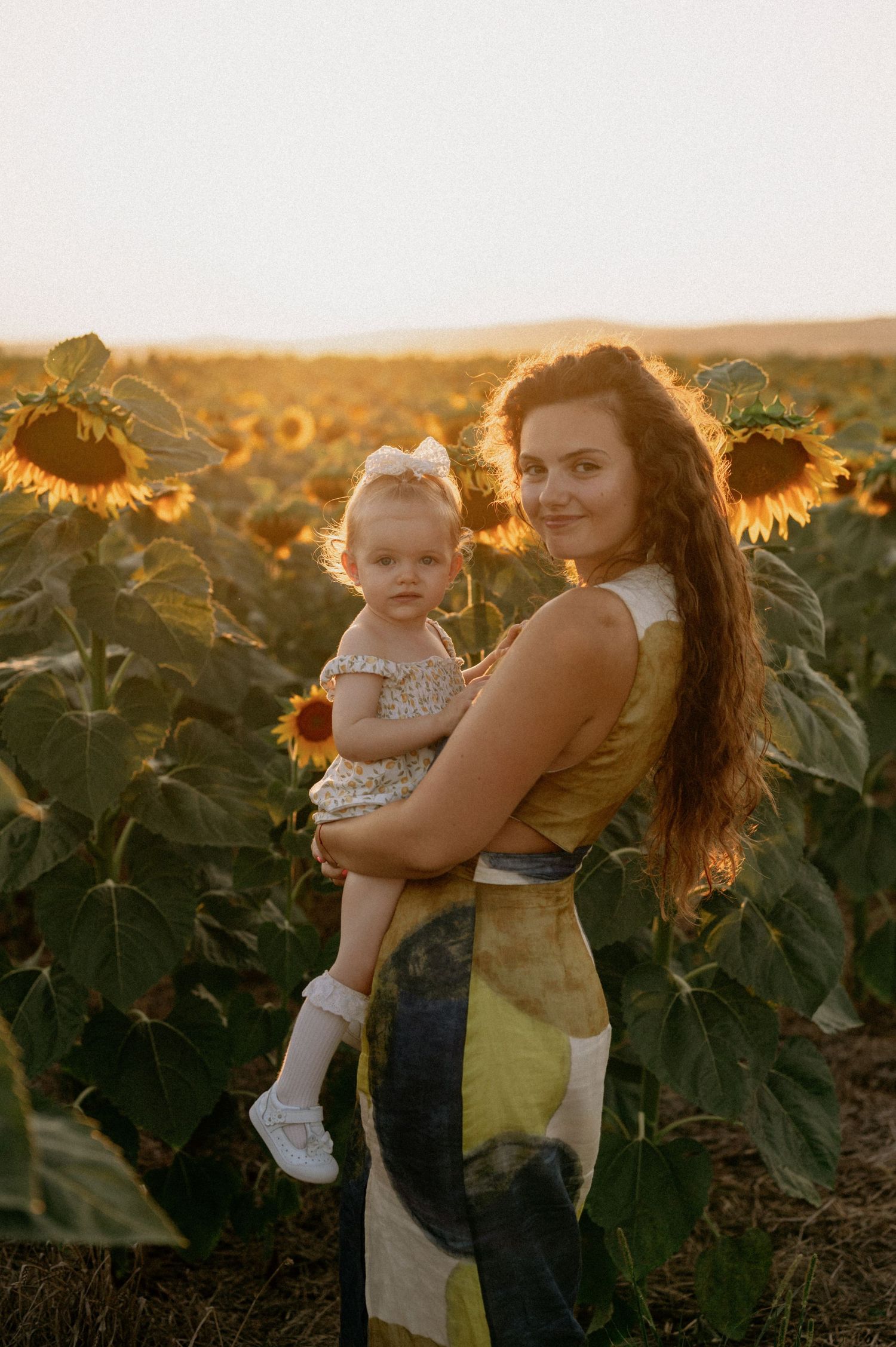 Woman in yellow dress holds toddler girl in sunflower field at golden hour sunset.