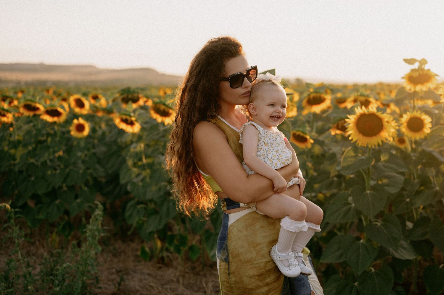 A mother holds her smiling baby in a sunflower field at golden hour sunset.