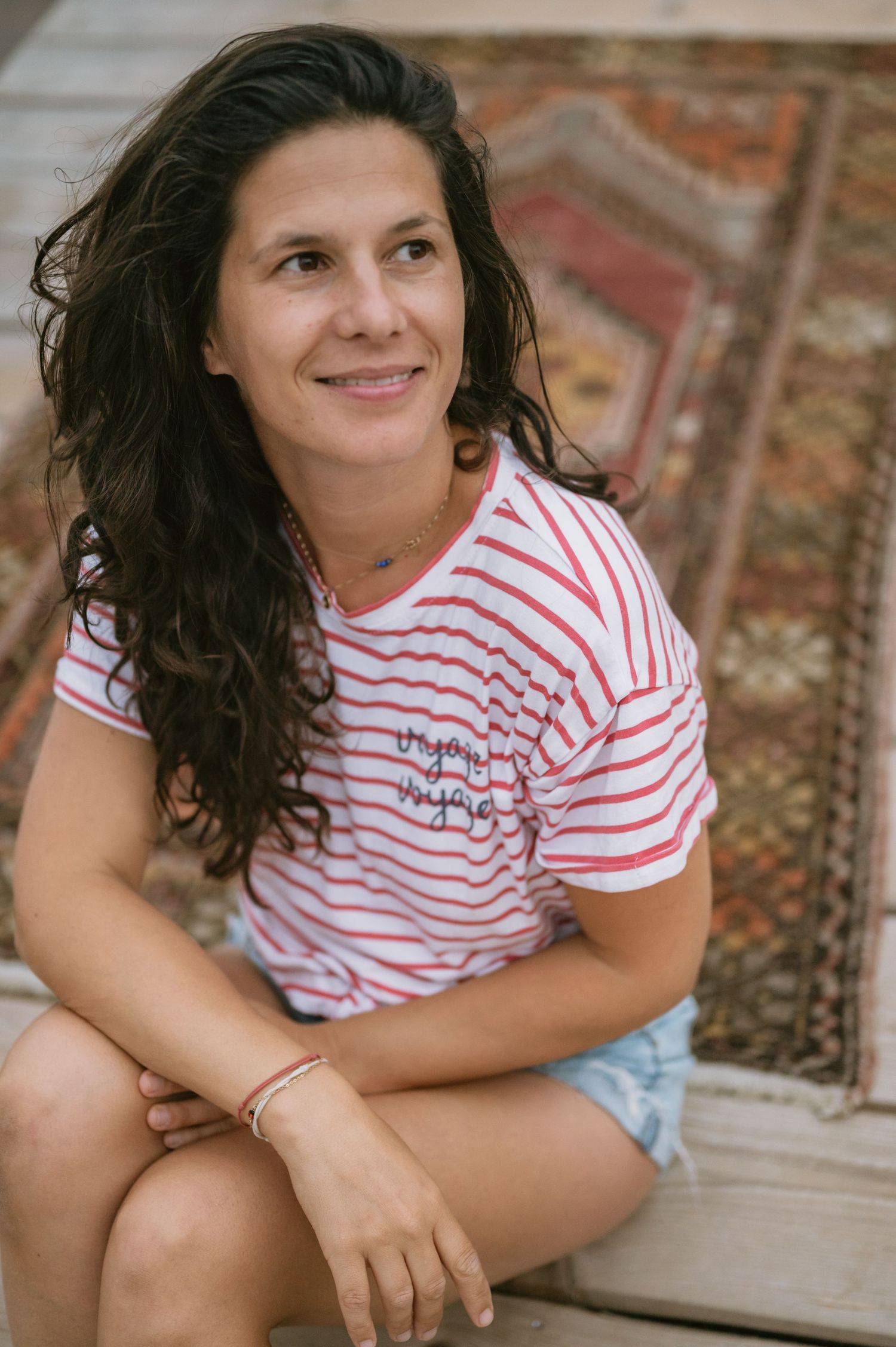 A smiling woman with curly dark hair sits outdoors wearing a red striped t-shirt and denim shorts, with a colorful rug behind her.