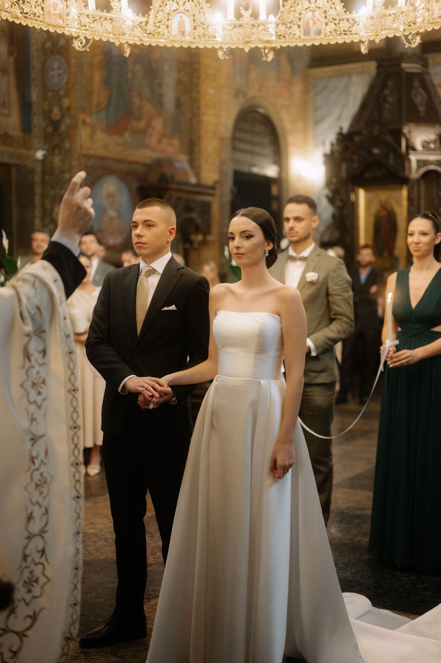 An elegant Orthodox church wedding ceremony with a couple in formal attire under ornate chandeliers and religious icons.