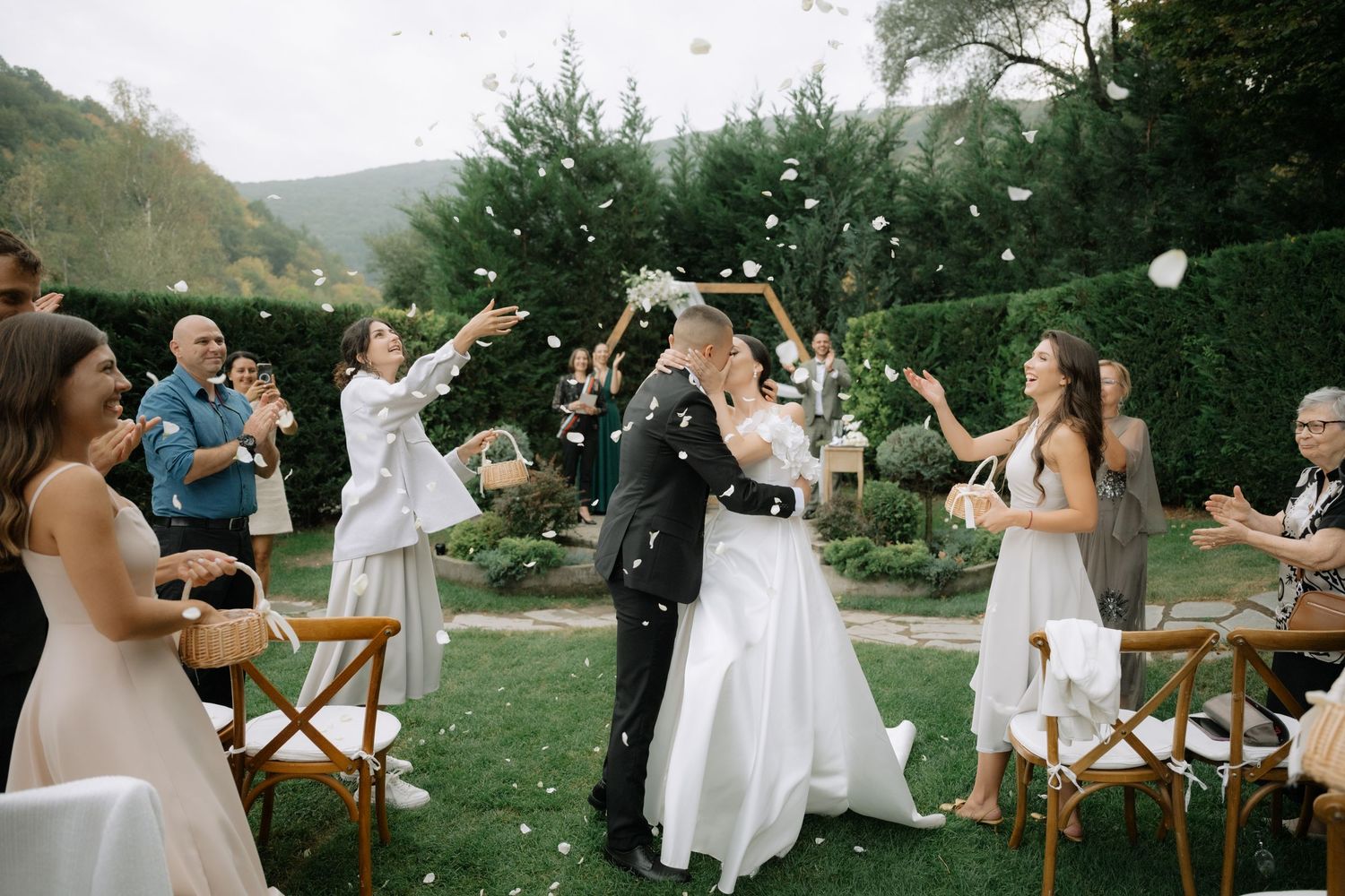 Wedding guests toss flower petals and celebrate during an outdoor garden ceremony at sunset.