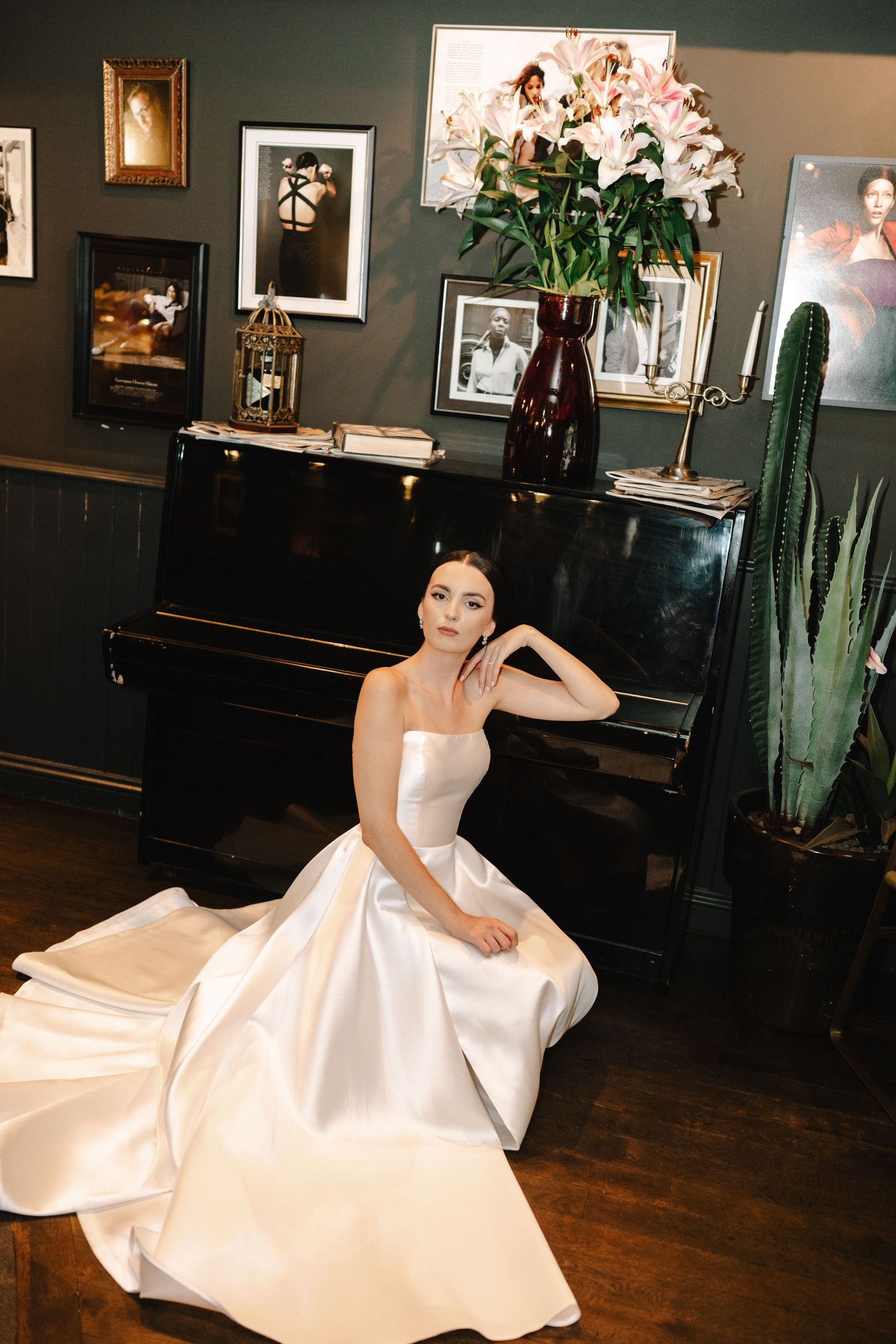 A model in an elegant white satin gown poses dramatically on a dark hardwood floor in a gallery-style room.