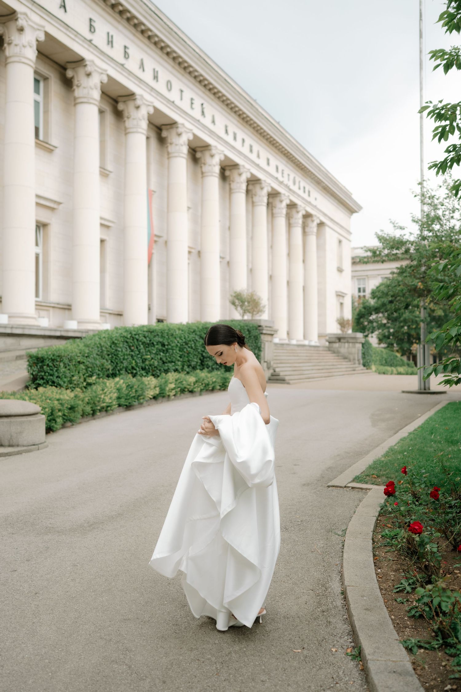 A bride in a flowing white wedding dress walks along a curved path outside a grand neoclassical building with tall columns.