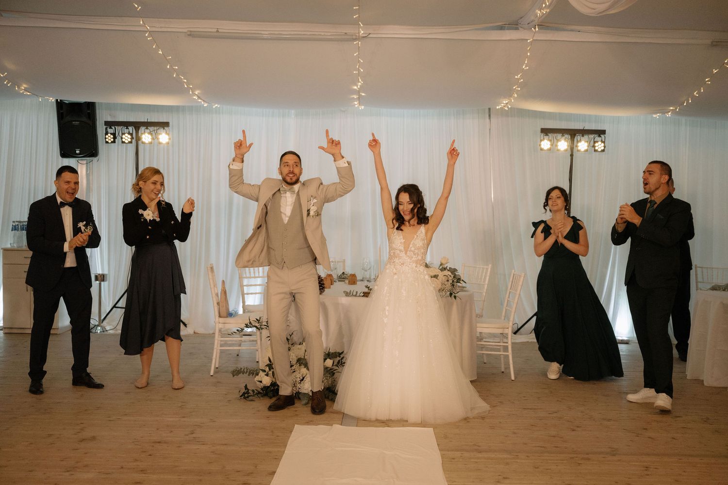 Bride and groom celebrate with raised arms at wedding reception, surrounded by guests cheering in elegant white decorated venue.