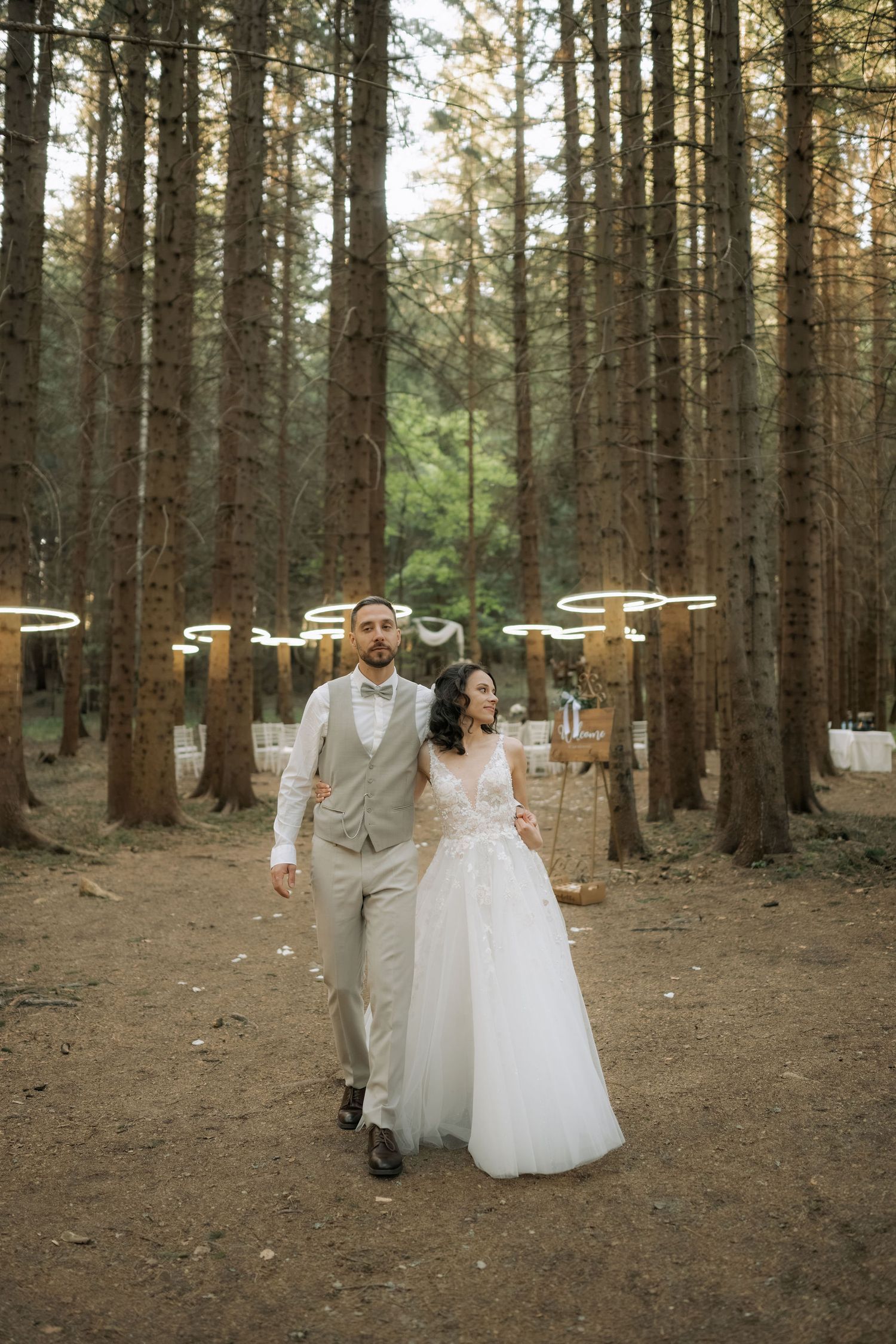 Bride and groom walking hand in hand through a forest wedding venue with glowing circular lights among tall pine trees.