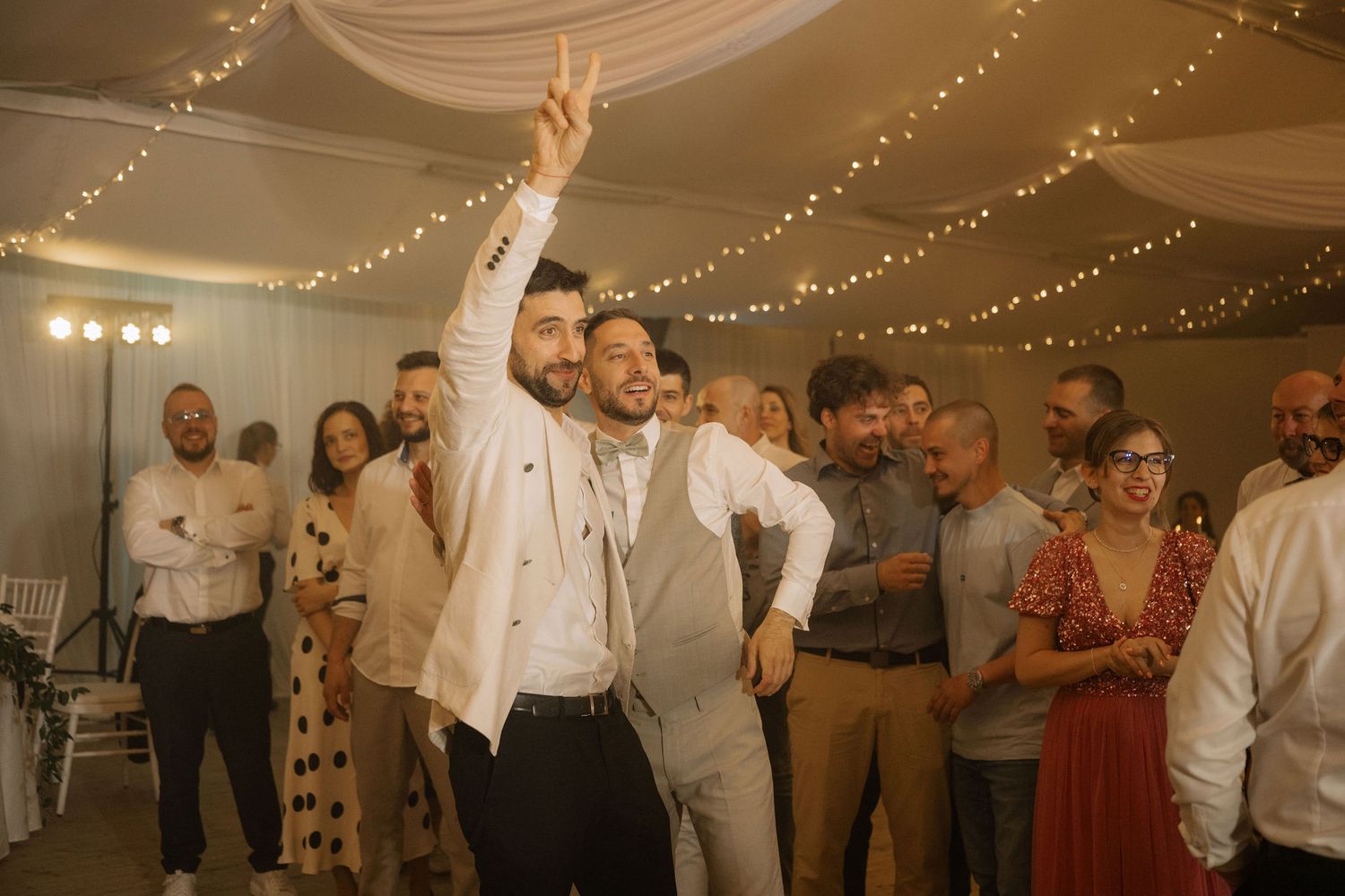 Two men dancing joyfully at a wedding reception under fairy lights with guests celebrating in the background.