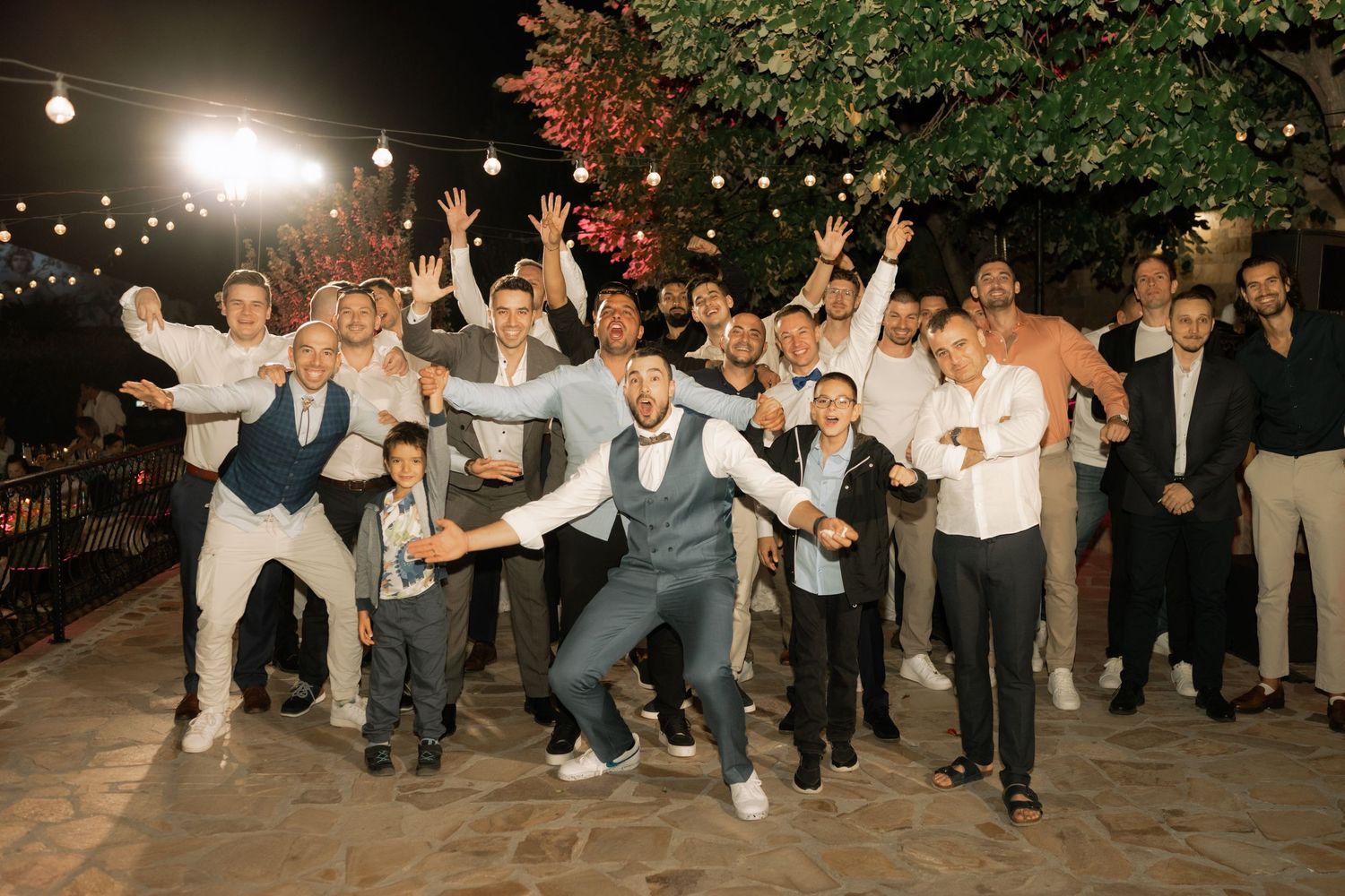 A celebratory group poses energetically on an outdoor patio at night under string lights during a festive event.