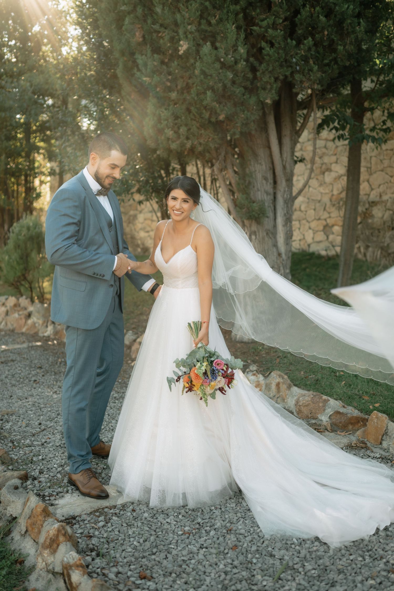 A wedding couple poses outdoors with a flowing white veil catching the light in a romantic woodland setting.