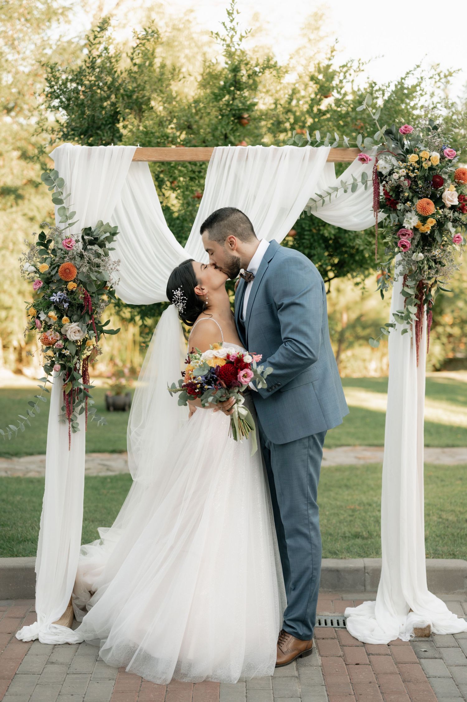 A romantic wedding ceremony under a white draped arch decorated with pink and red floral arrangements in an outdoor garden setting.