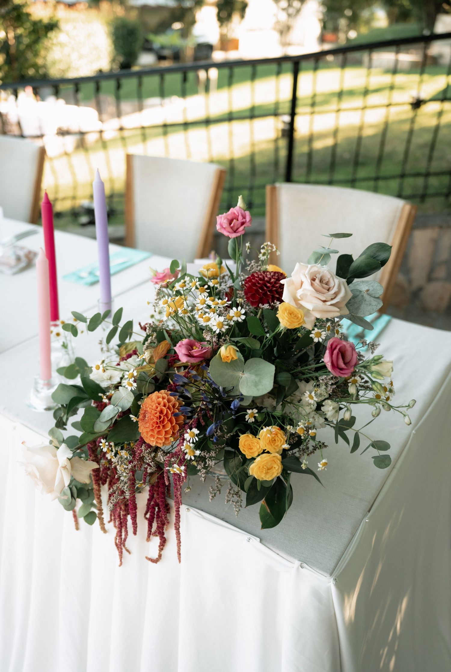 Elegant floral centerpiece with pink roses, yellow dahlias, and eucalyptus on a white tablecloth with tall candles.