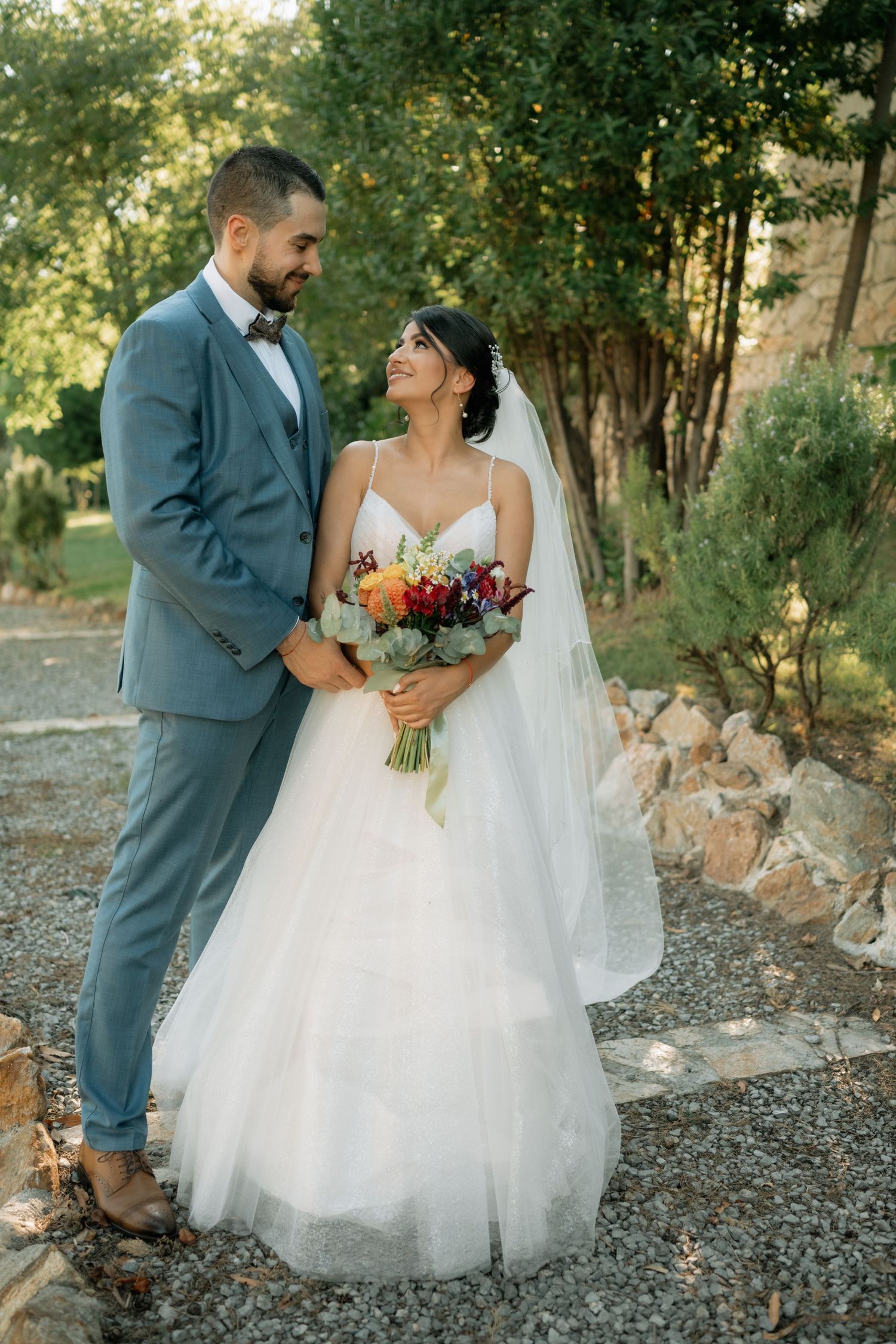 Bride and groom share intimate moments in a rustic outdoor setting with natural greenery and gravel path.