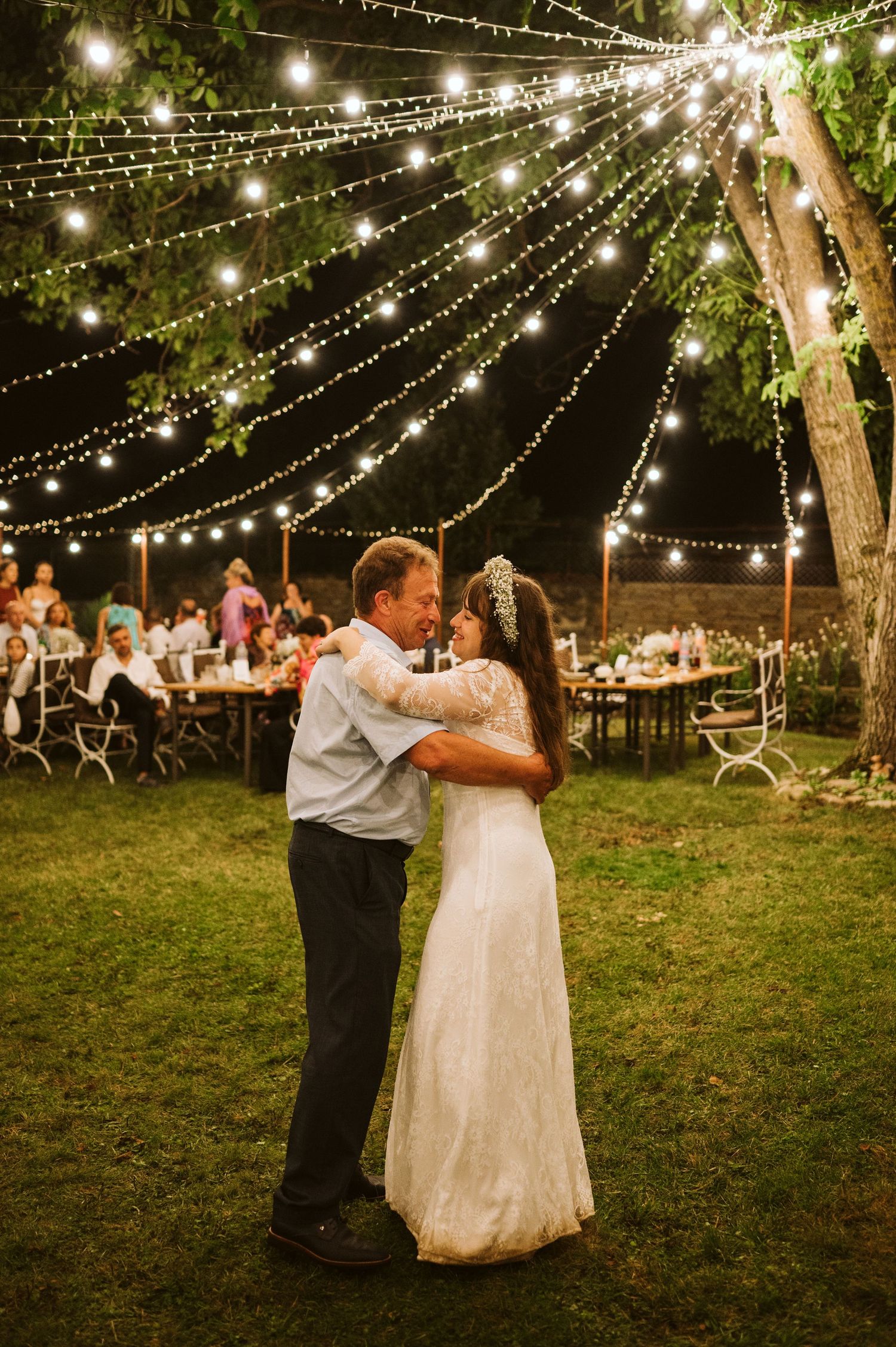 Newlyweds dance under twinkling string lights at an outdoor evening reception.