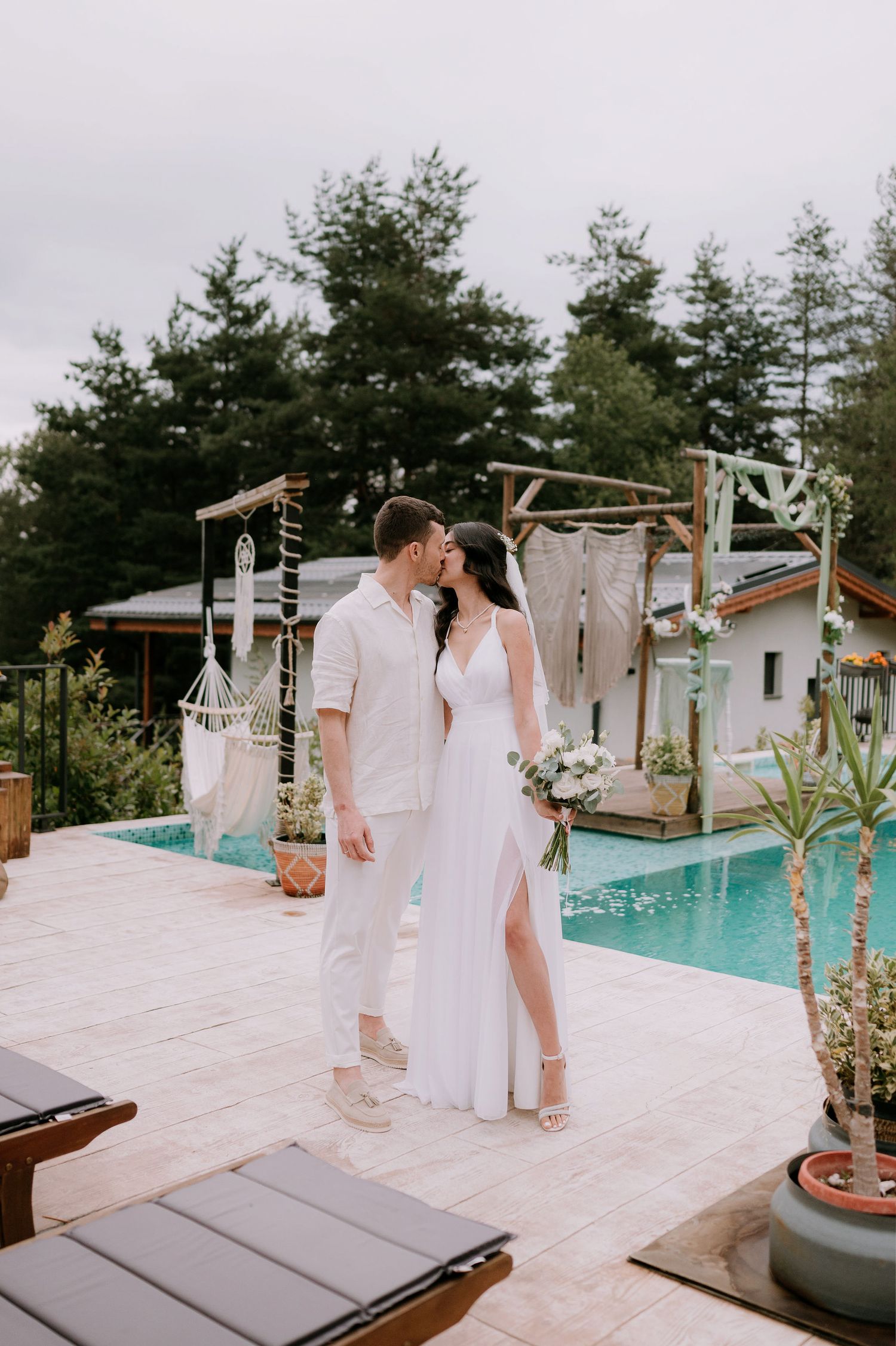 Elegant couple in white outfits pose for wedding photos near a swimming pool surrounded by potted plants.
