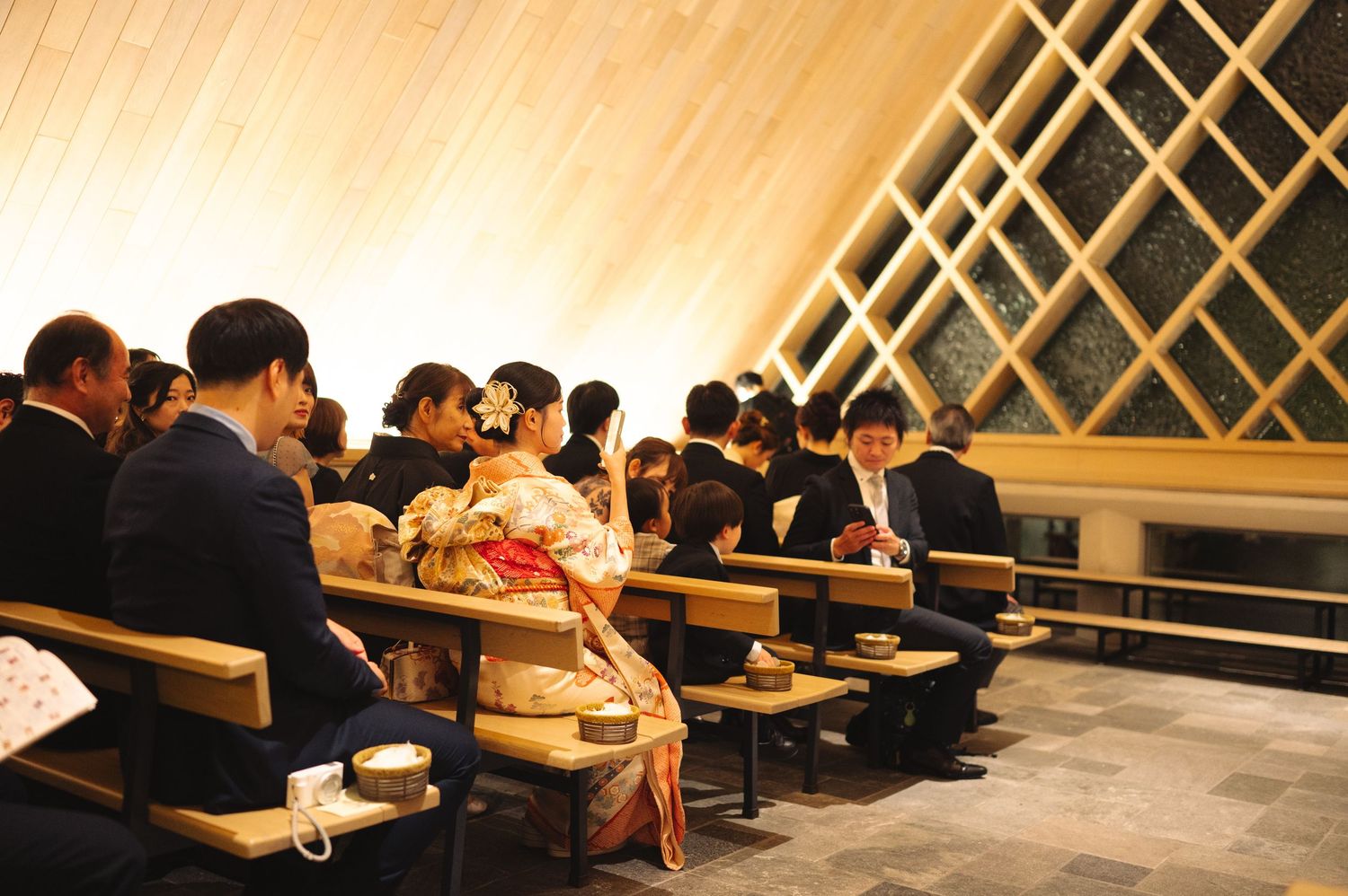 Audience members in formal wear seated in rows at venue with distinctive wooden lattice wall.