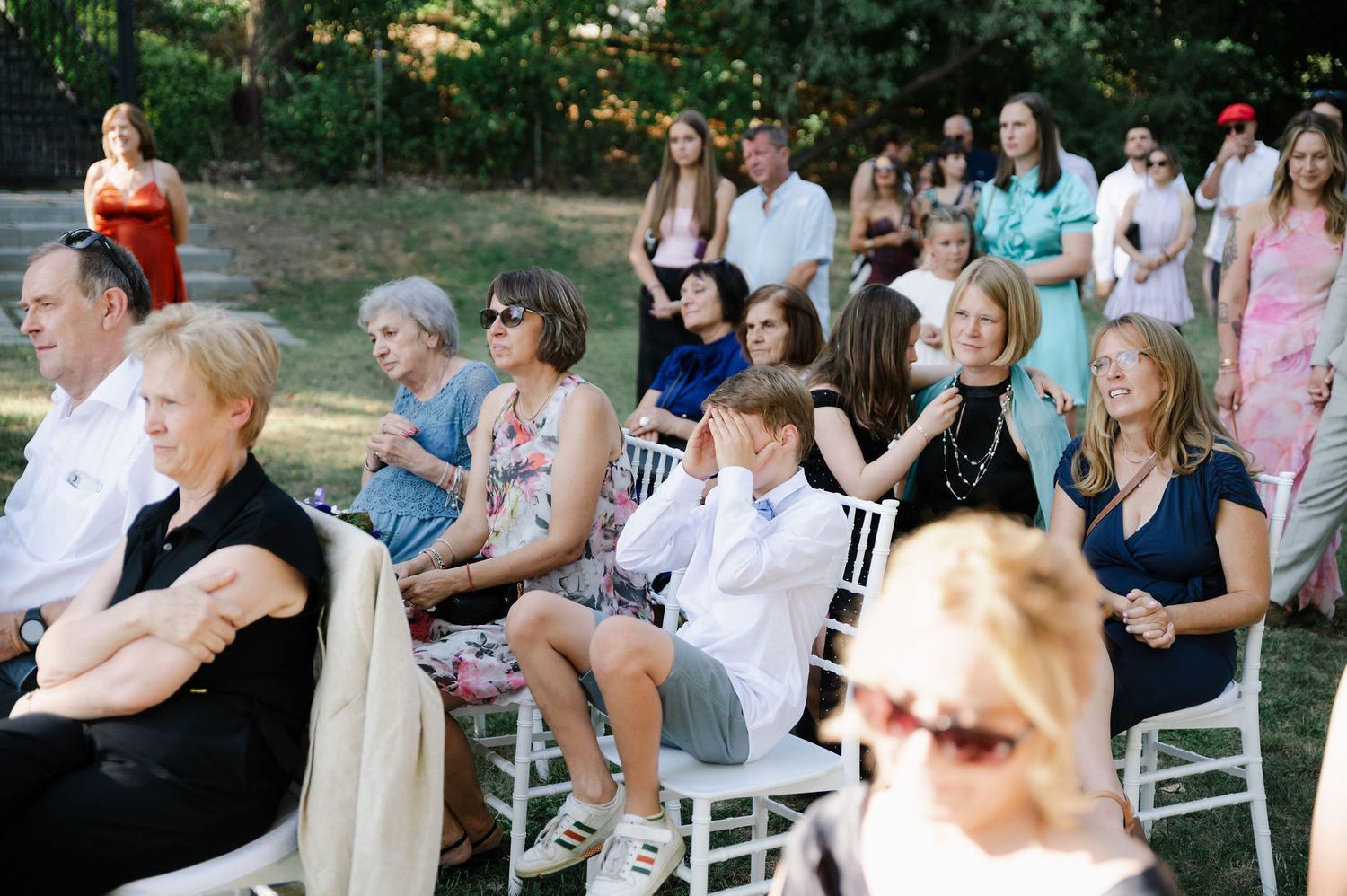 Wedding guests seated in white chairs watch ceremony outdoors.