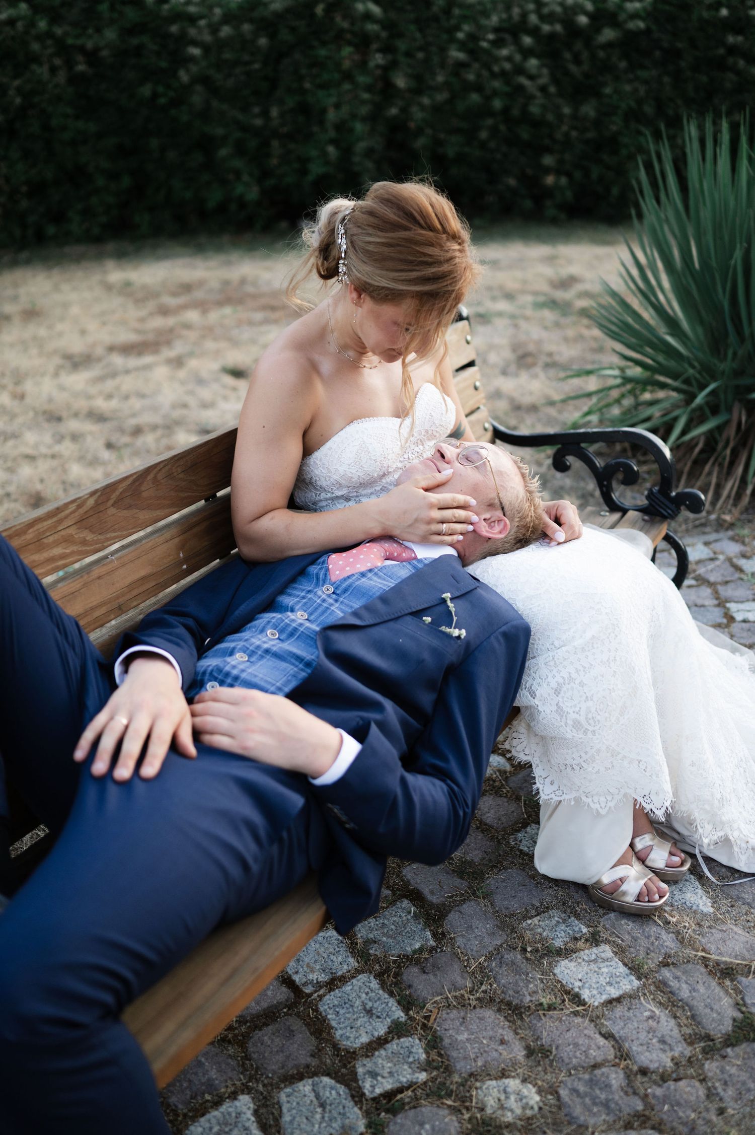 A couple in wedding attire embraces on a stone path bench during an outdoor photoshoot.