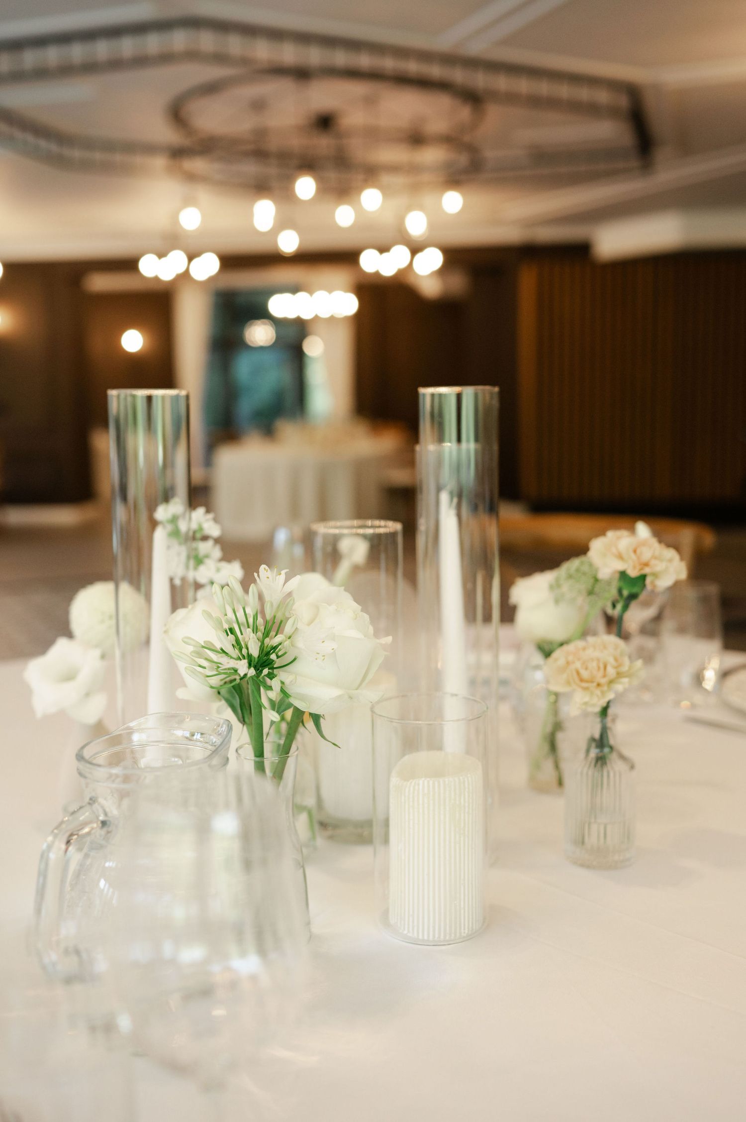 Modern wedding table decorations with glass cylinder vases and white flowers under warm chandelier lighting.
