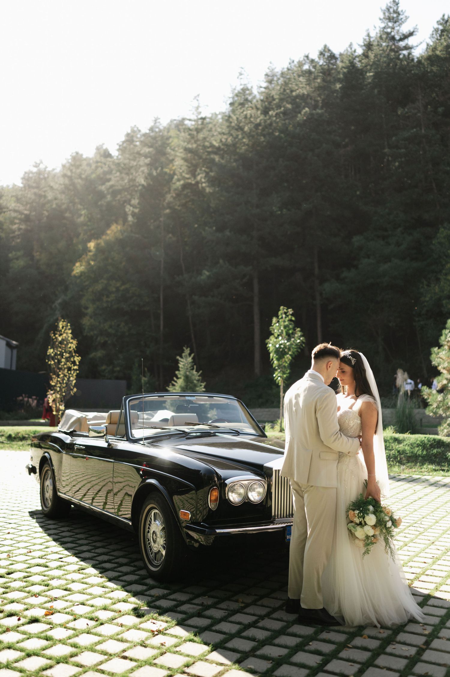 Sunlit wedding portrait with a classic black convertible on cobblestone pavement.