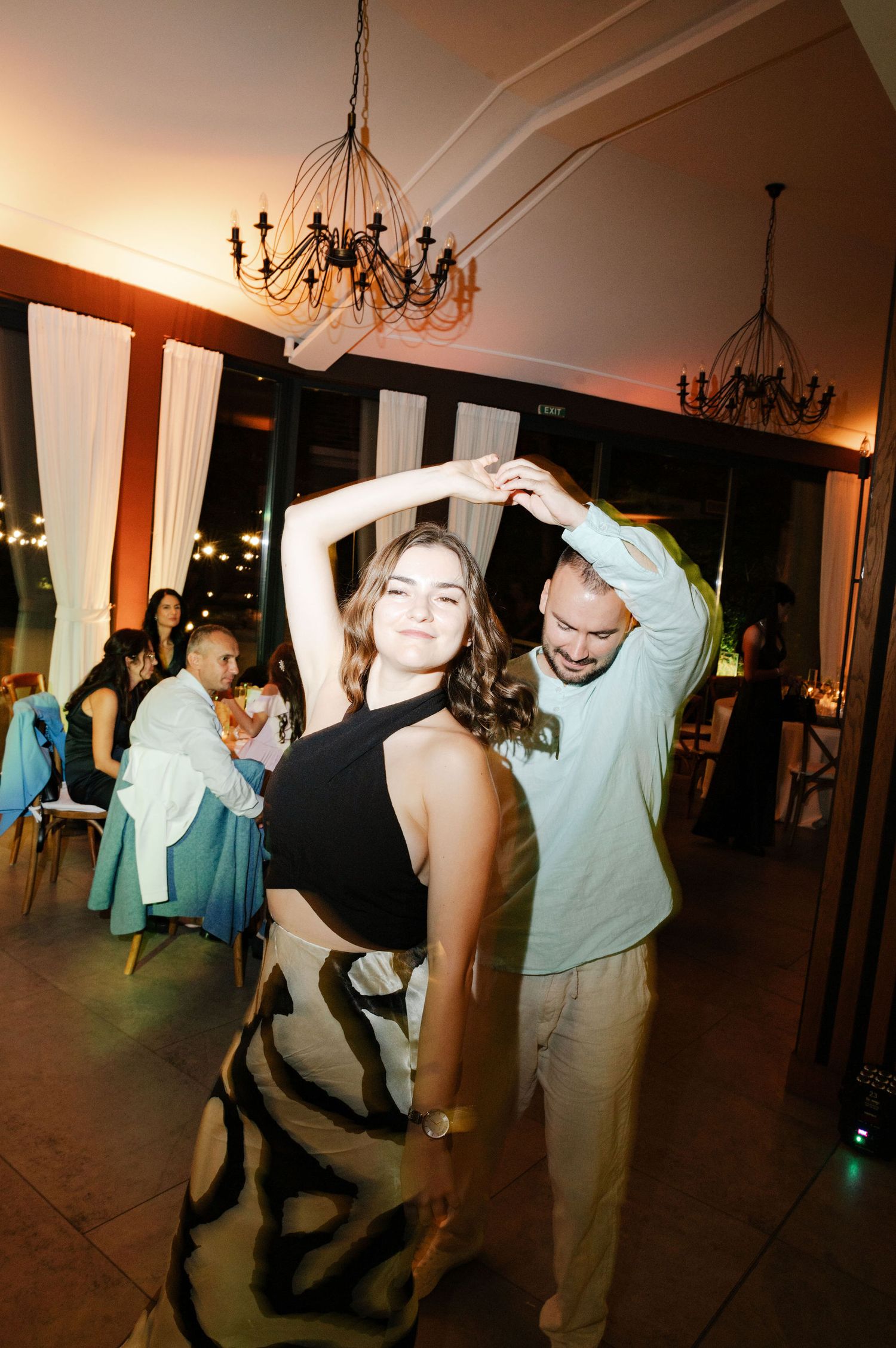 Wedding party guests dancing under elegant chandeliers in warm ambient lighting.